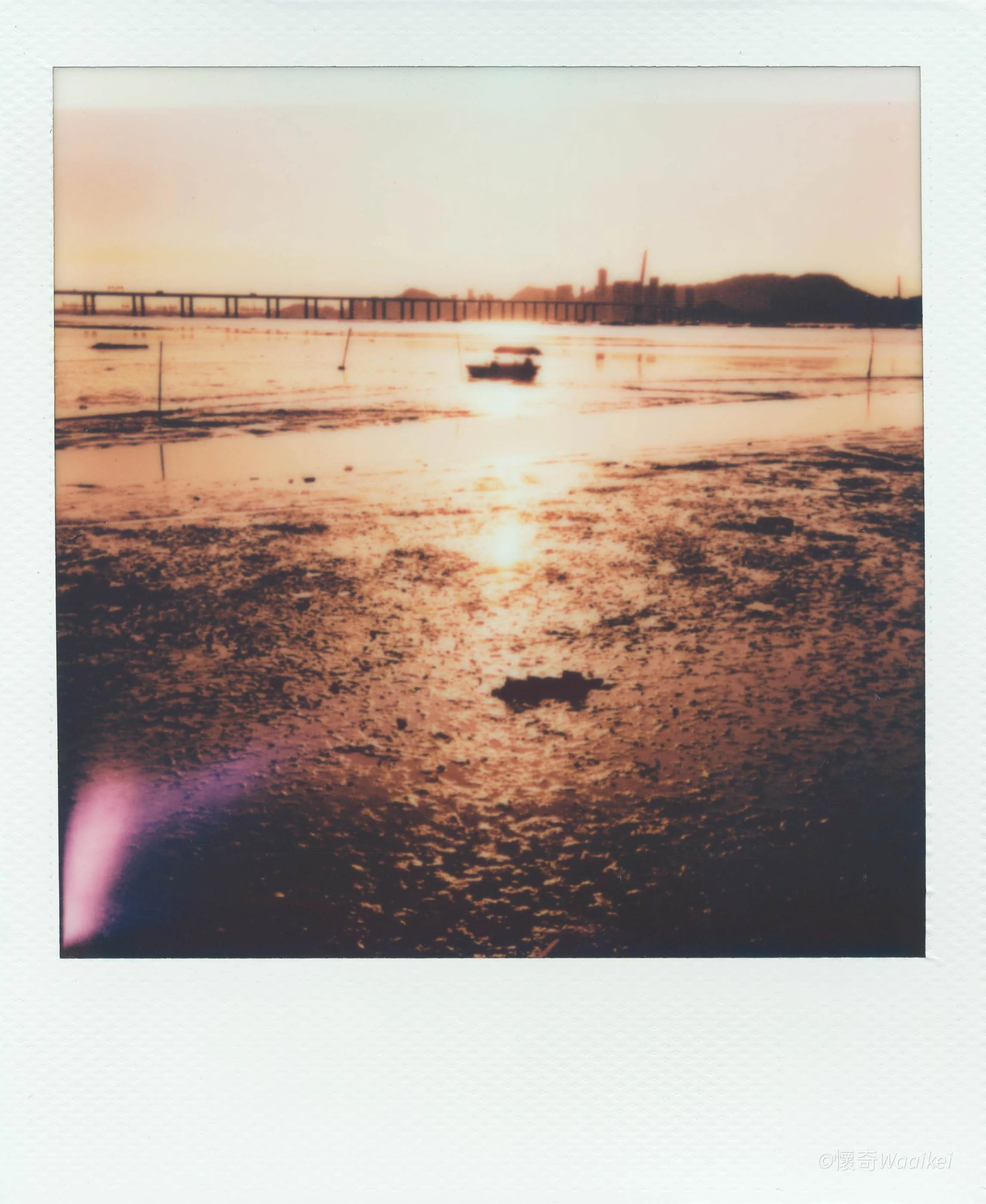 Polaroid picture of a sunset over a body of water with a boat and a city skyline in the background.