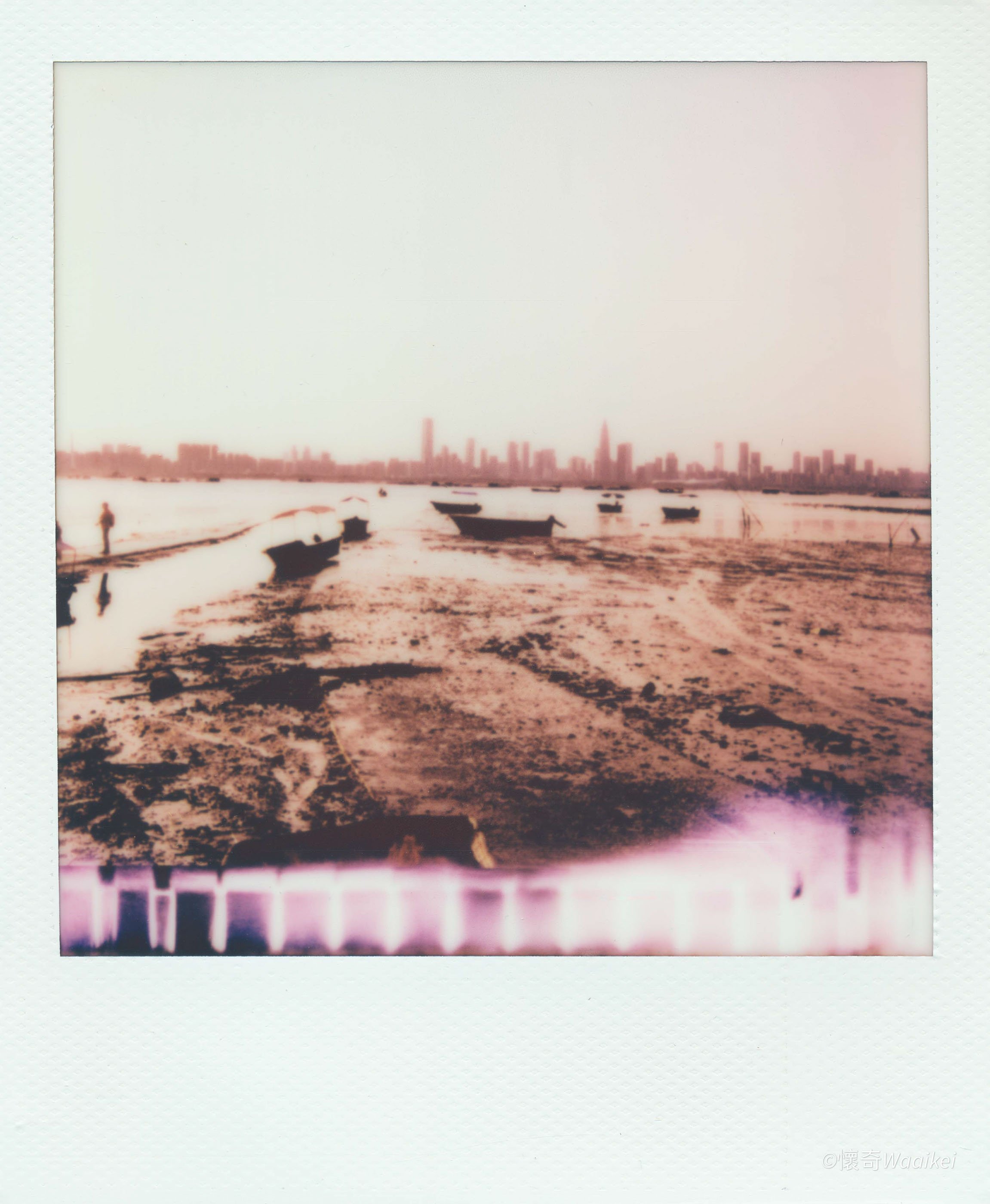 A Polaroid photo showing a beach with boats and a distant city skyline with tall buildings, under an overcast sky.