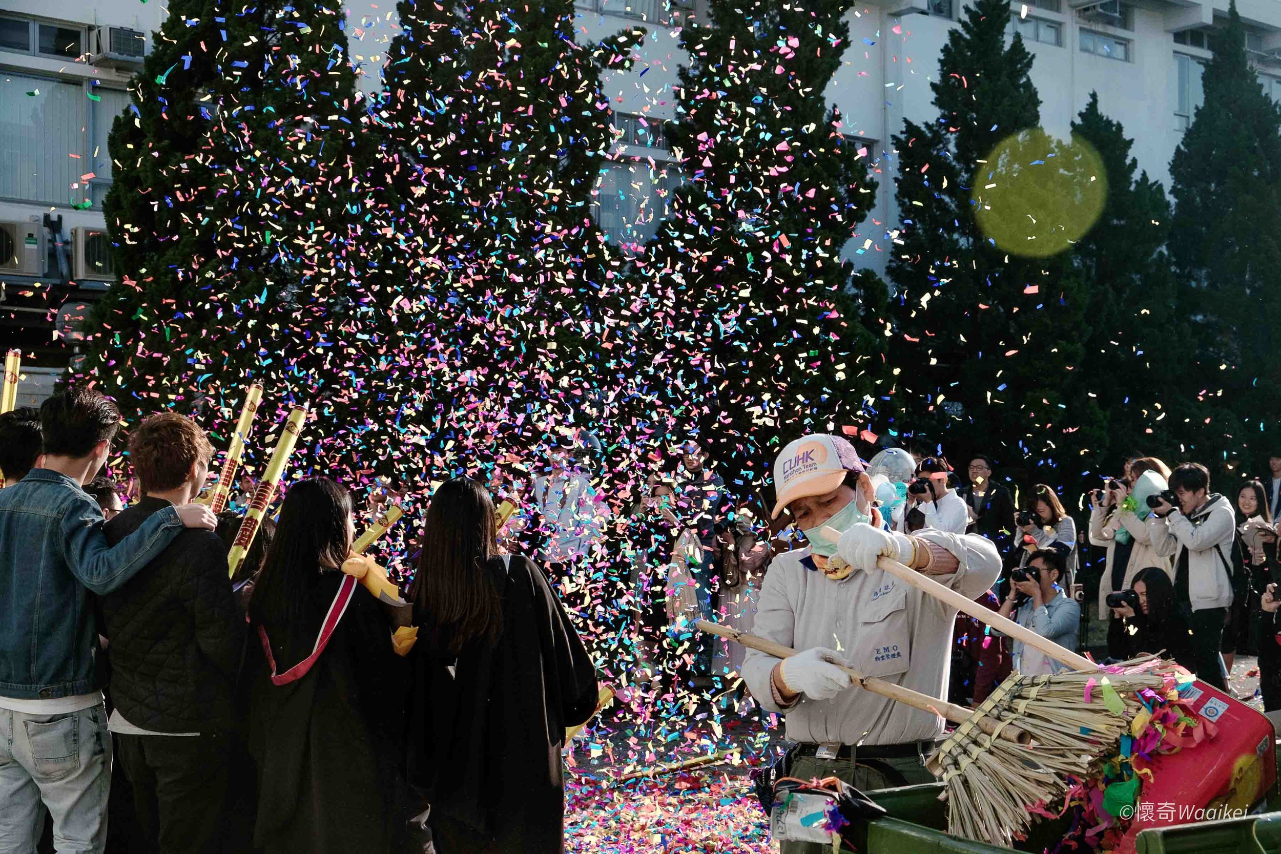 People gathered outdoors during a celebration, with a man operating fireworks or firecrackers, and colorful confetti falling through the air. Tall trees and a building are visible in the background.