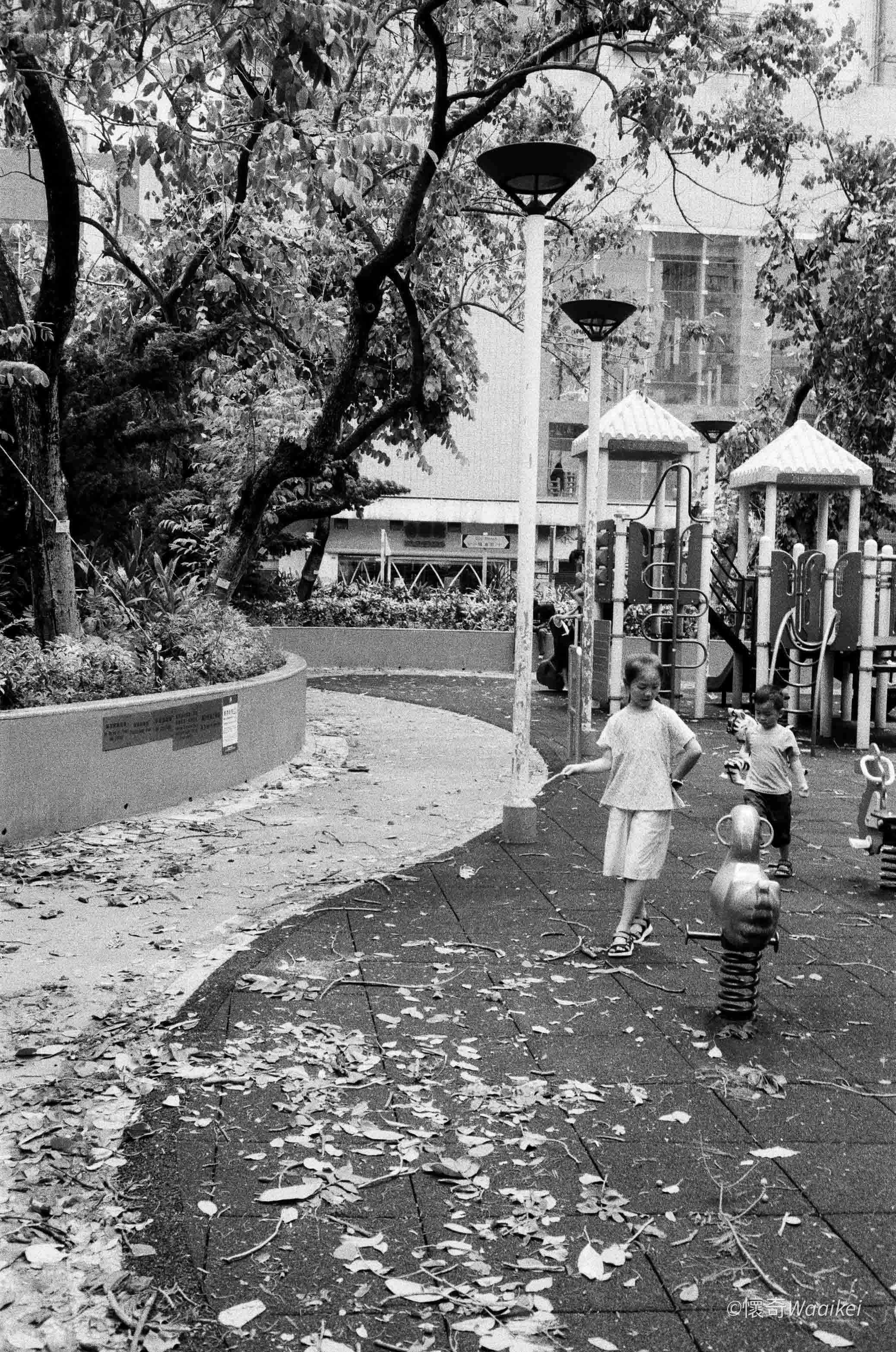 A black and white photo of children playing in a park with fallen leaves on the ground, playground equipment, and trees in the background.