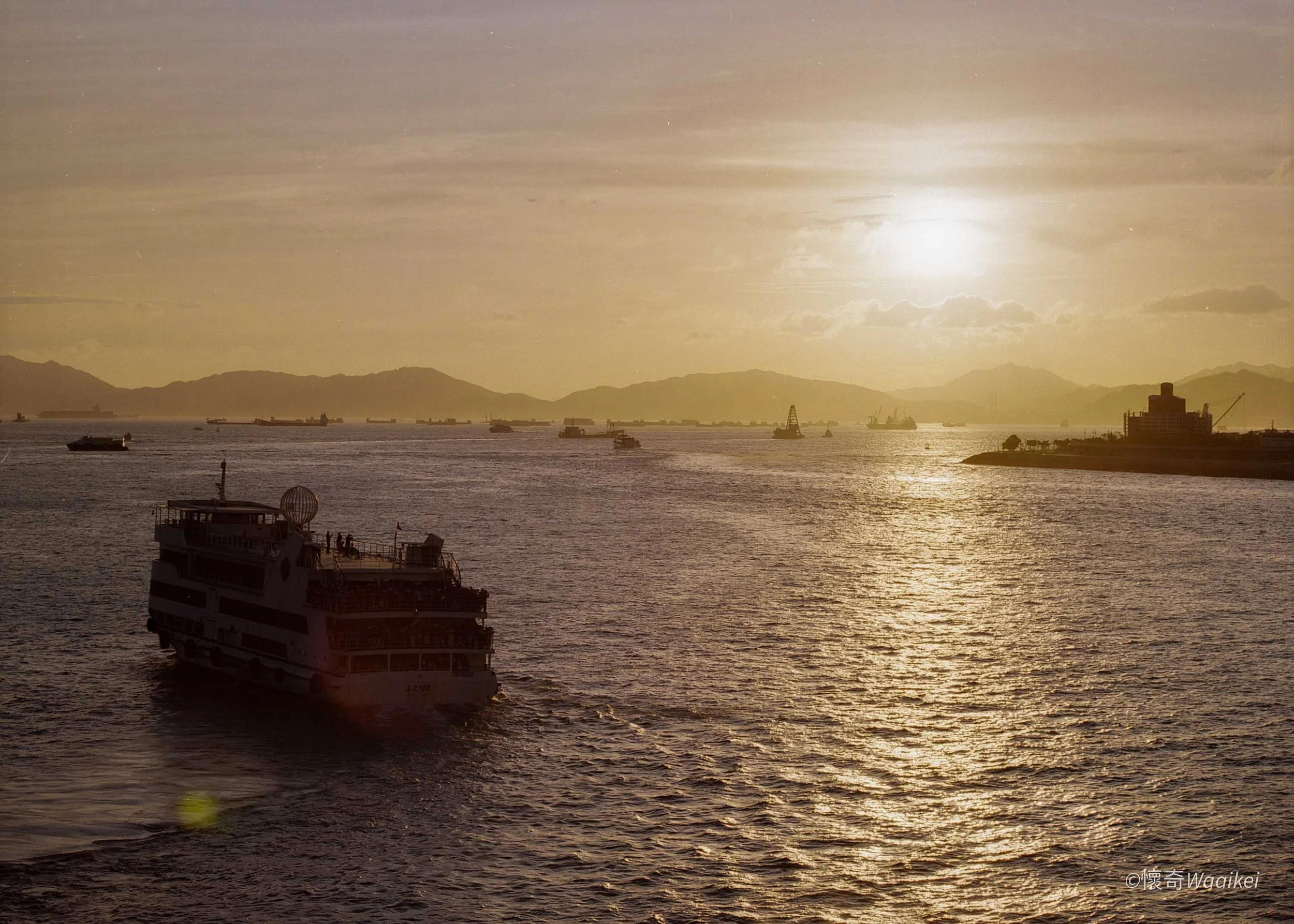Sunset over a body of water with several boats and ships, mountains in the background, and a large cruise ship in the foreground.