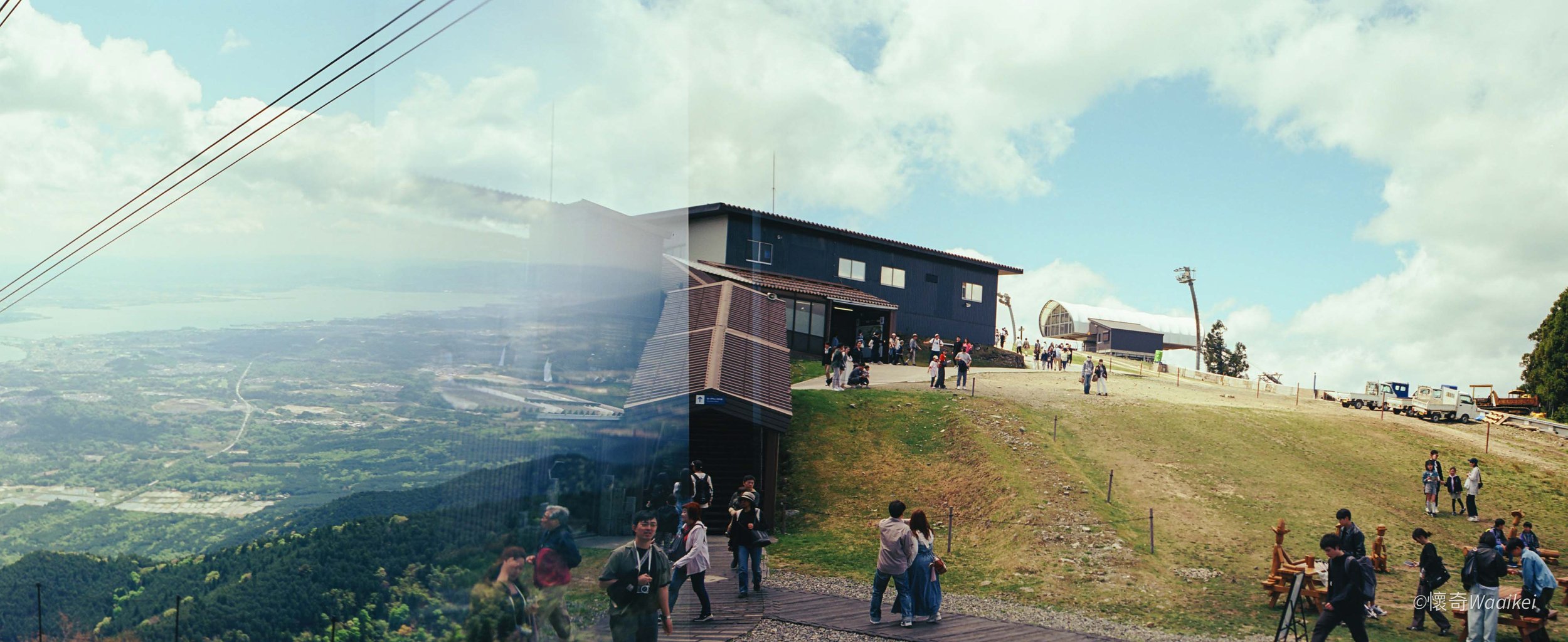 People gathering outside a modern building on a hilltop with scenic views, a grassy incline, and equipment structures in the background under a partly cloudy sky.