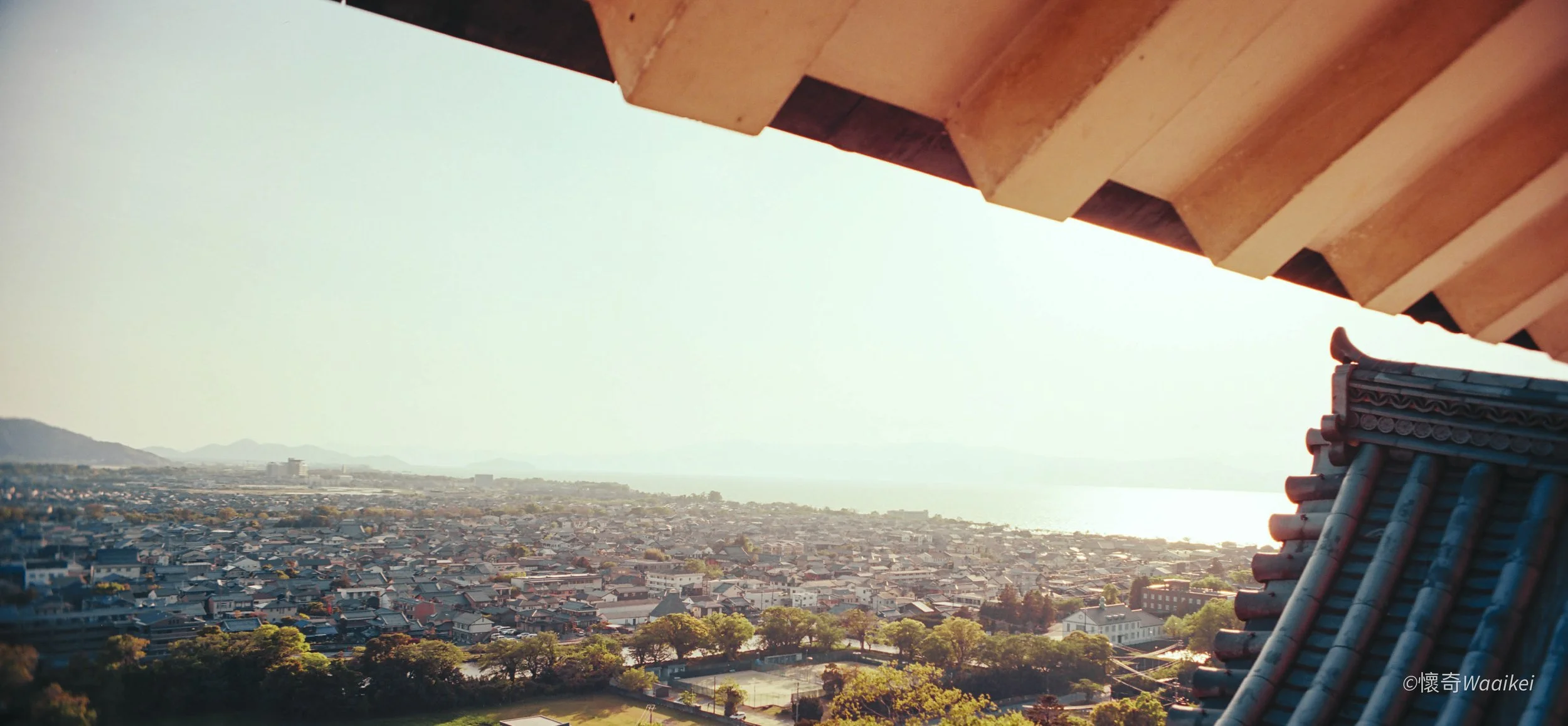 An aerial view of a cityscape with a mix of traditional and modern buildings, body of water in the distance, and mountains on the horizon, seen from a high vantage point underneath a wooden roof.