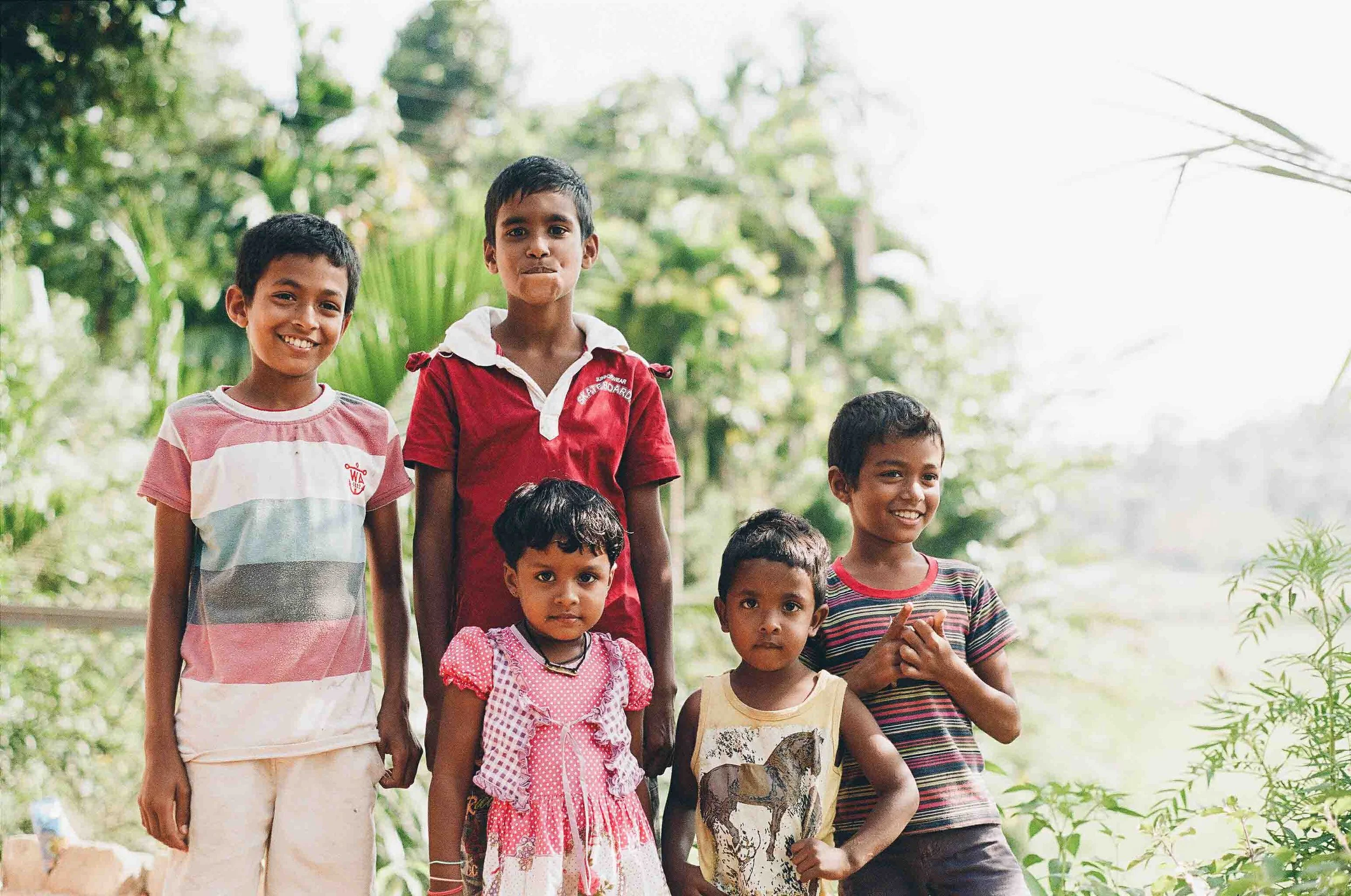 Group of seven children outdoors, some smiling and some looking serious, surrounded by greenery.