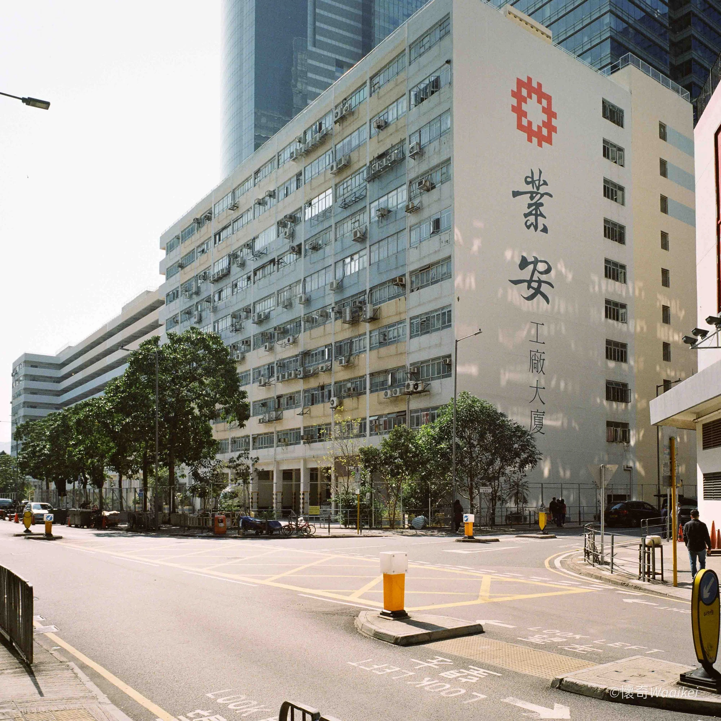 Urban street scene with tall buildings, trees, parked bicycles, sidewalk, and street signs, including a large building with Asian characters and a red geometric logo