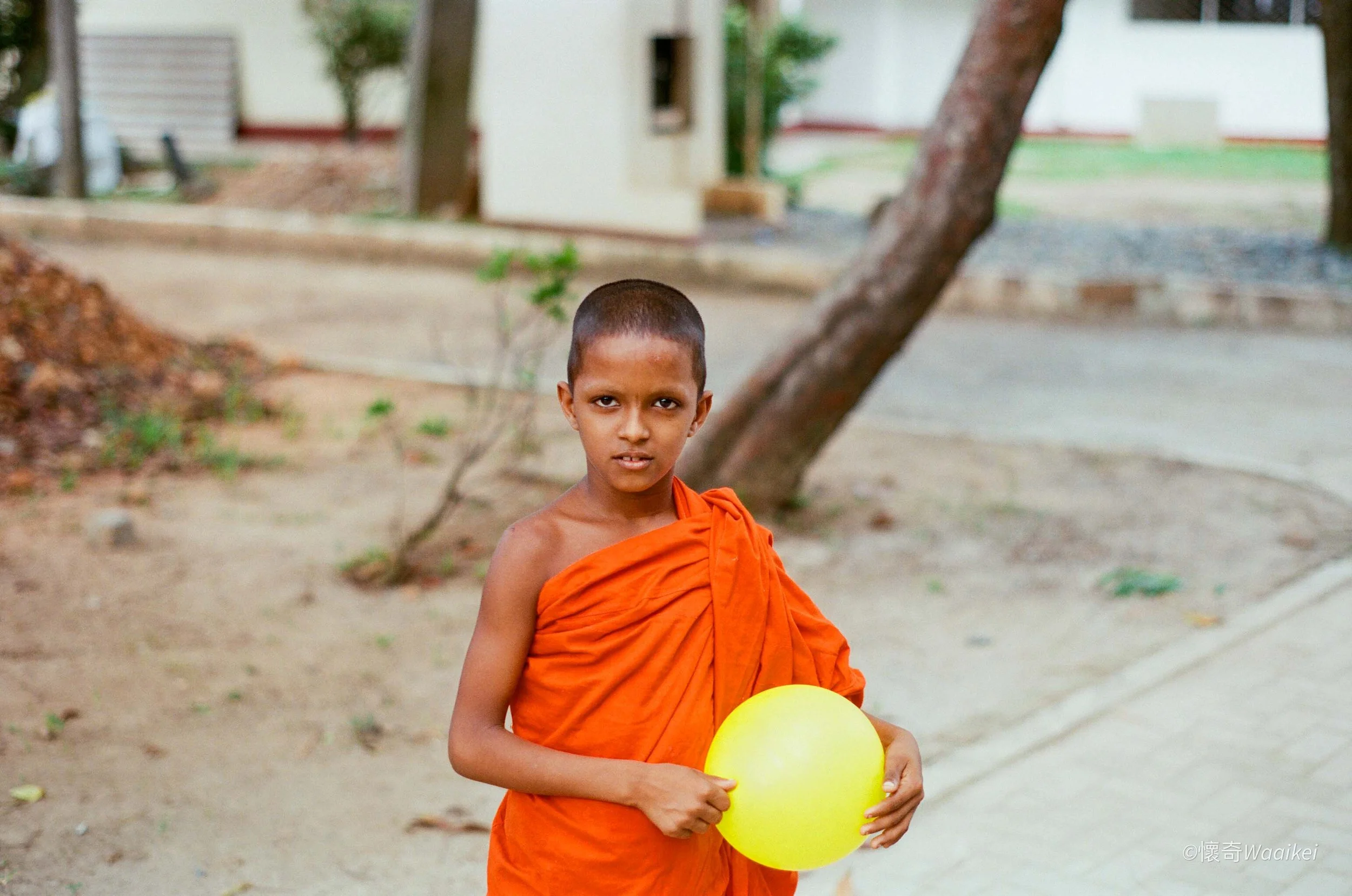 A young boy dressed as a monk in an orange robe holding yellow balloons outdoors in a natural setting.