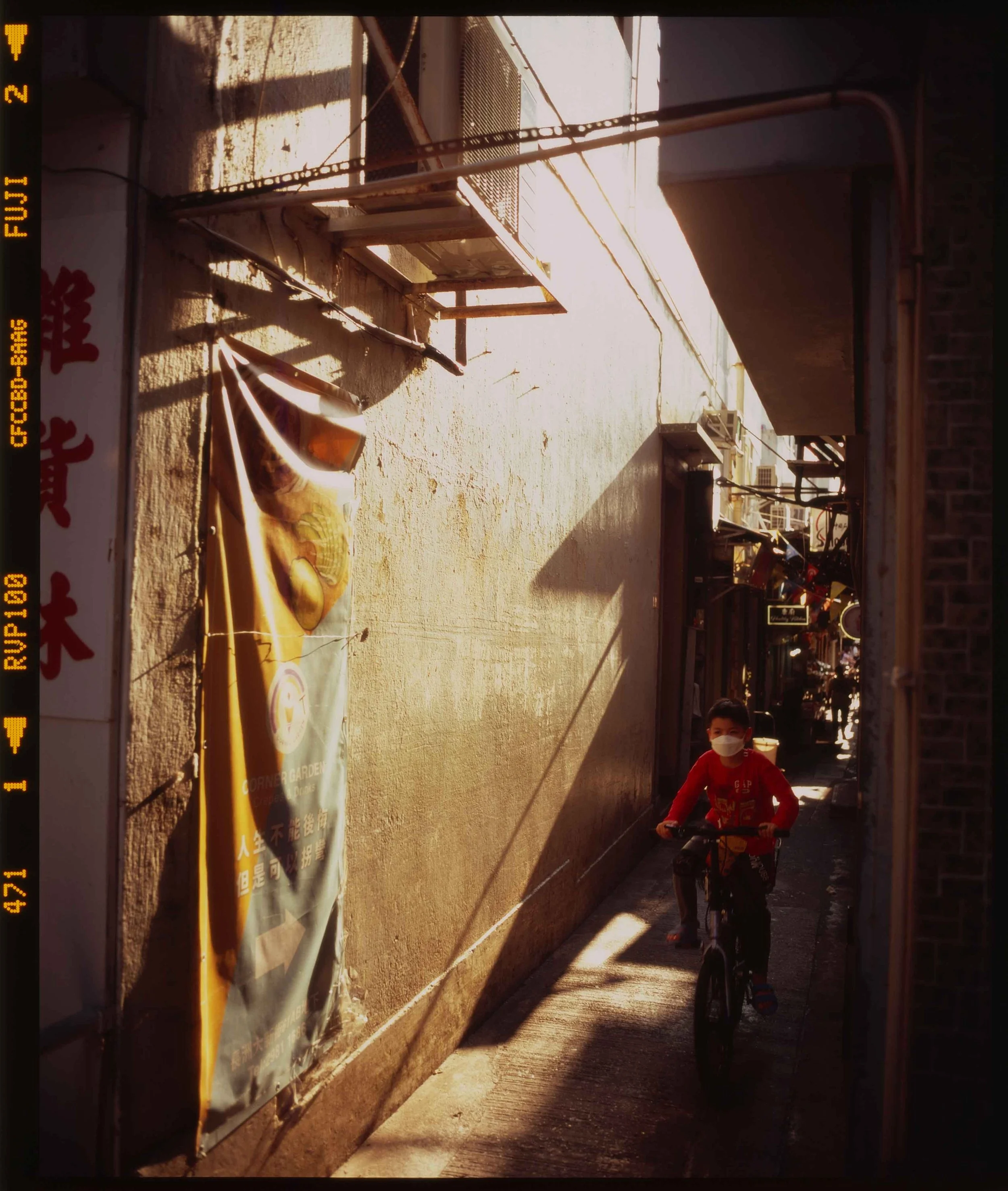 A young child riding a bicycle on a rooftop balcony during sunset, with shadows cast on the wall and a banner with Chinese characters on the left side.