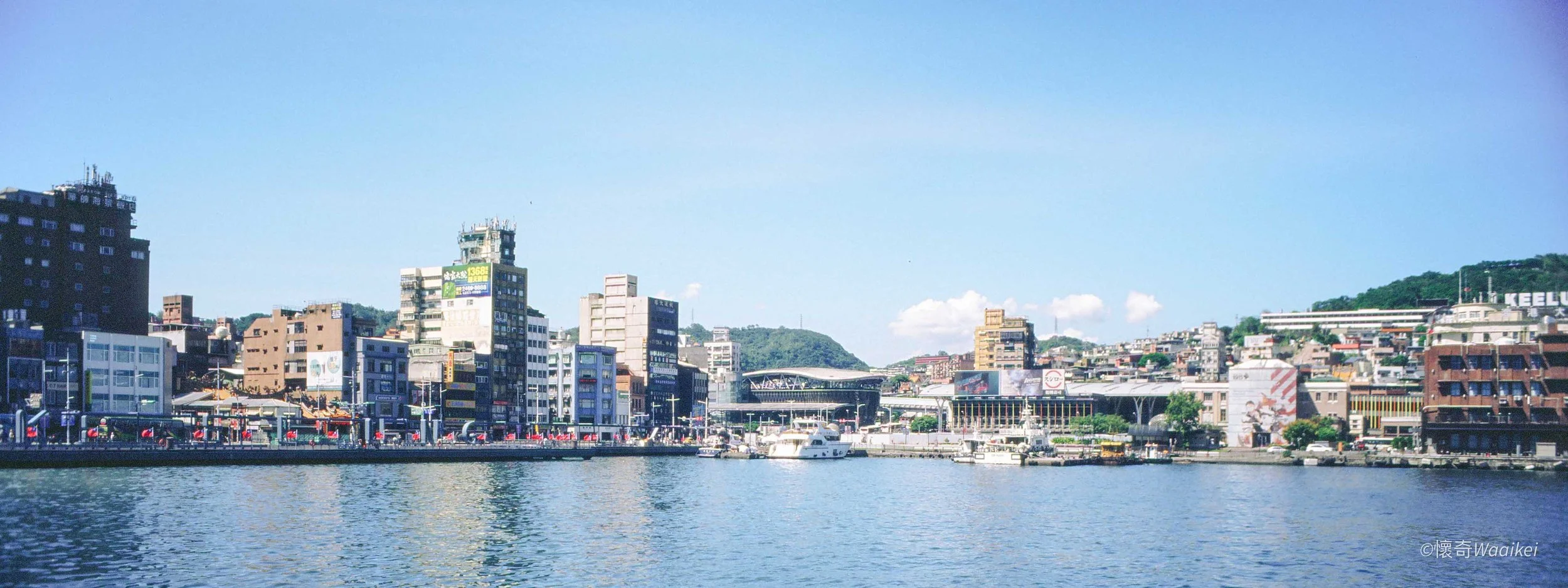Cityscape with waterfront, various buildings, a bridge, boats on the water, and a hillside with greenery in the background.