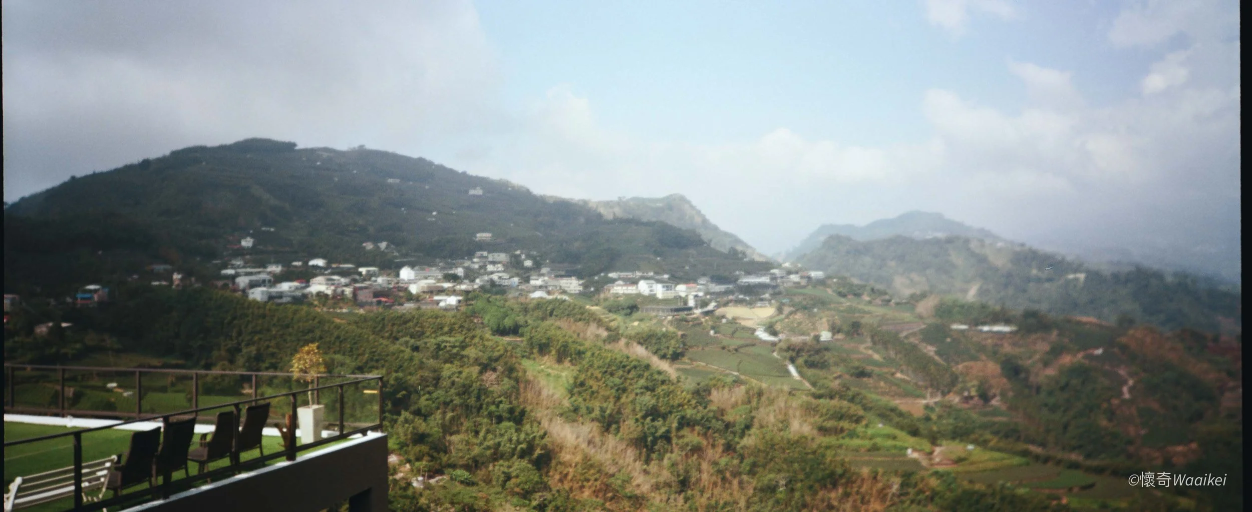 Scenic view of a mountainous landscape with a residential area and lush greenery, taken from a balcony with chairs and a railing in the foreground.