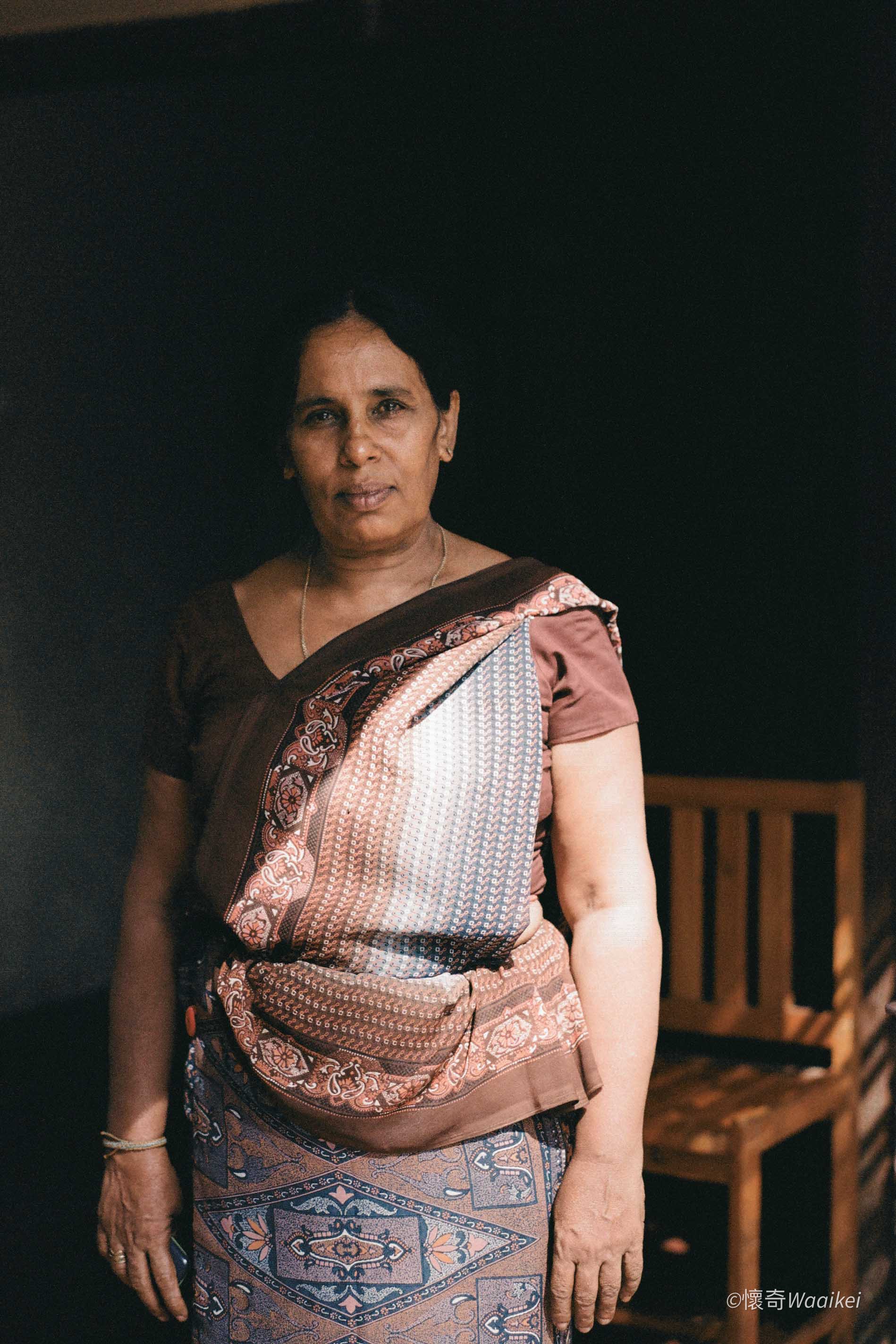 A woman with short black hair wearing a brown, pink, and white patterned traditional dress, standing in front of a dark background with a wooden structure visible on the side.