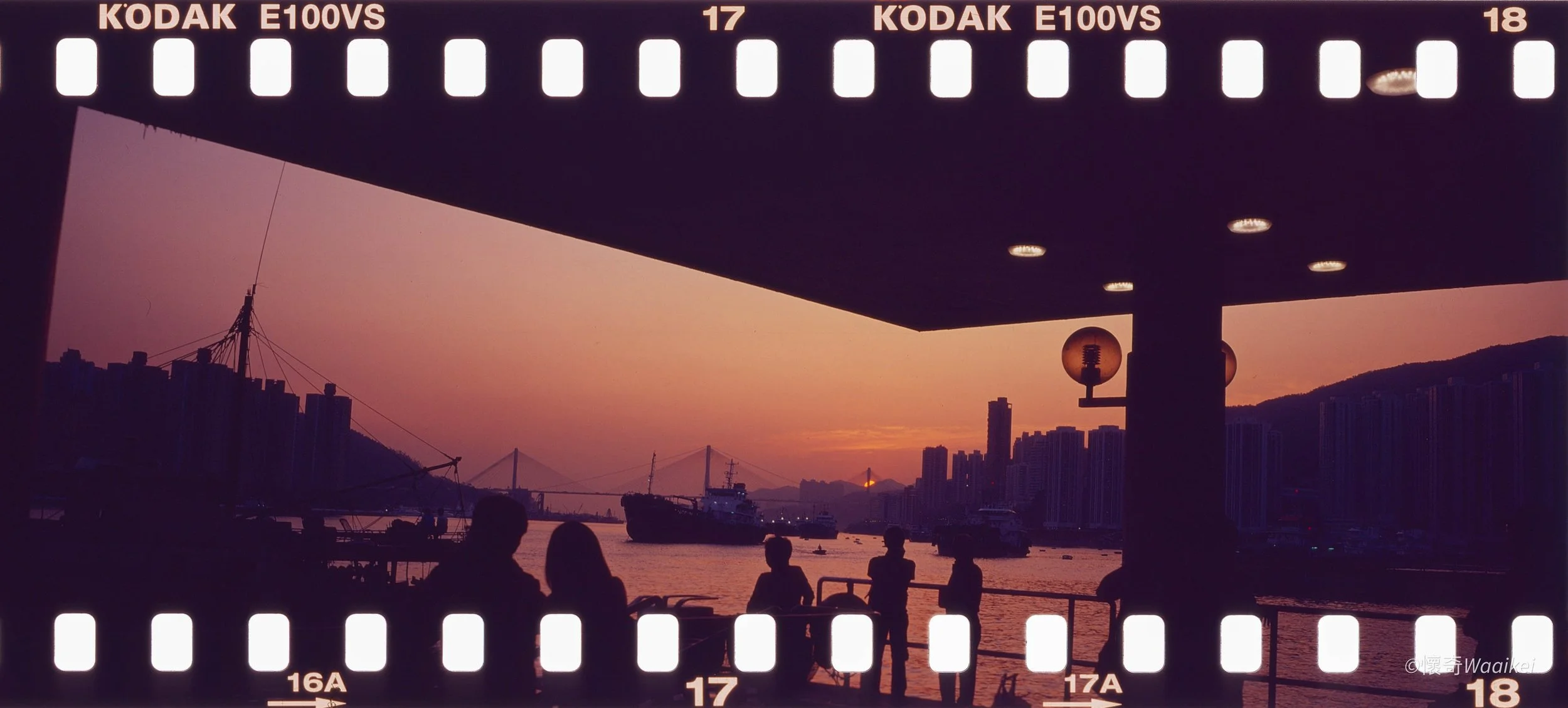 Silhouettes of people sitting and standing under a bridge at sunset with boats and city skyline in the background.