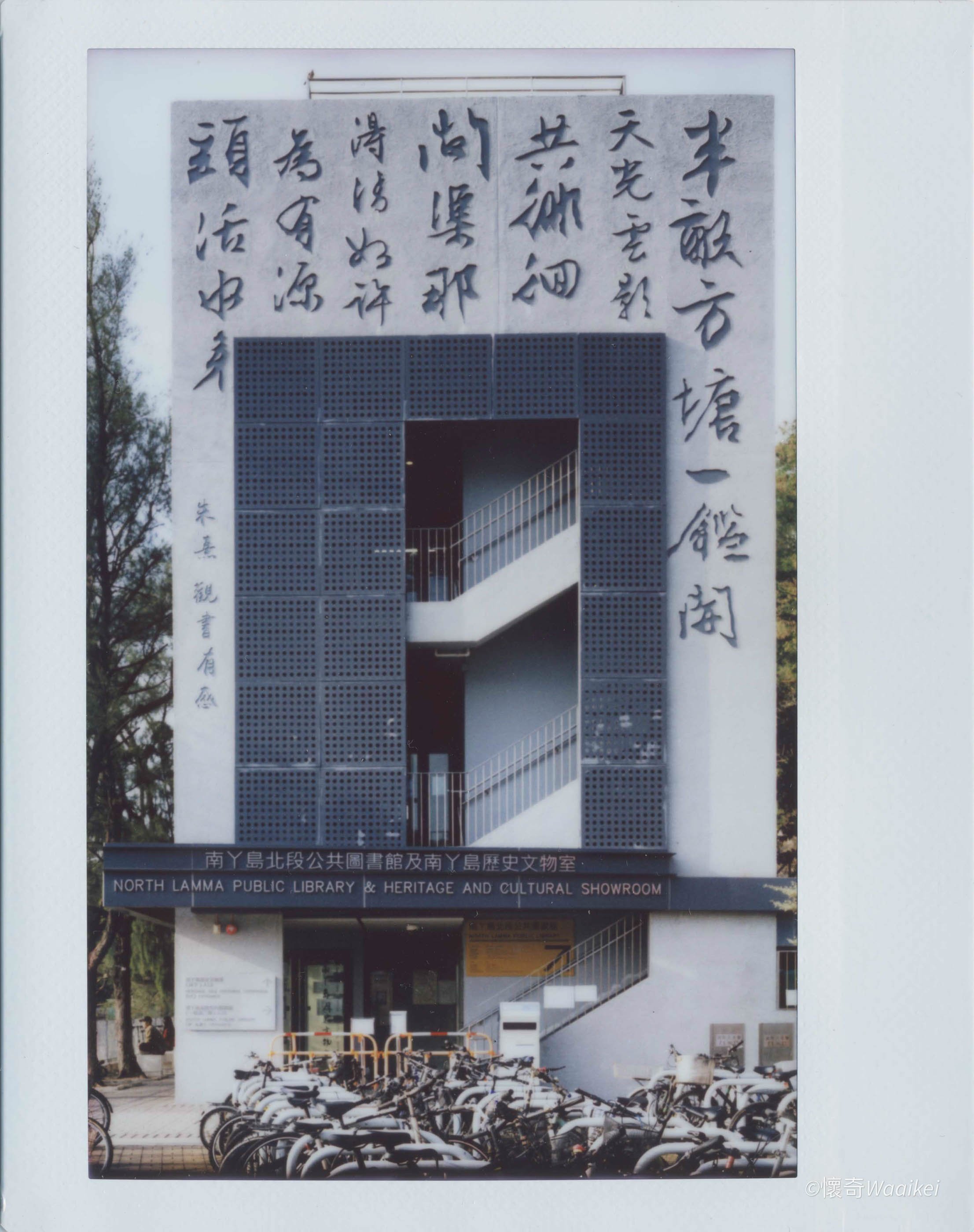 Photograph of the entrance to North Lamma Public Library and Heritage and Cultural Showroom, featuring a modern building with a large sign in Chinese and English, bicycles parked in front, and trees on either side.
