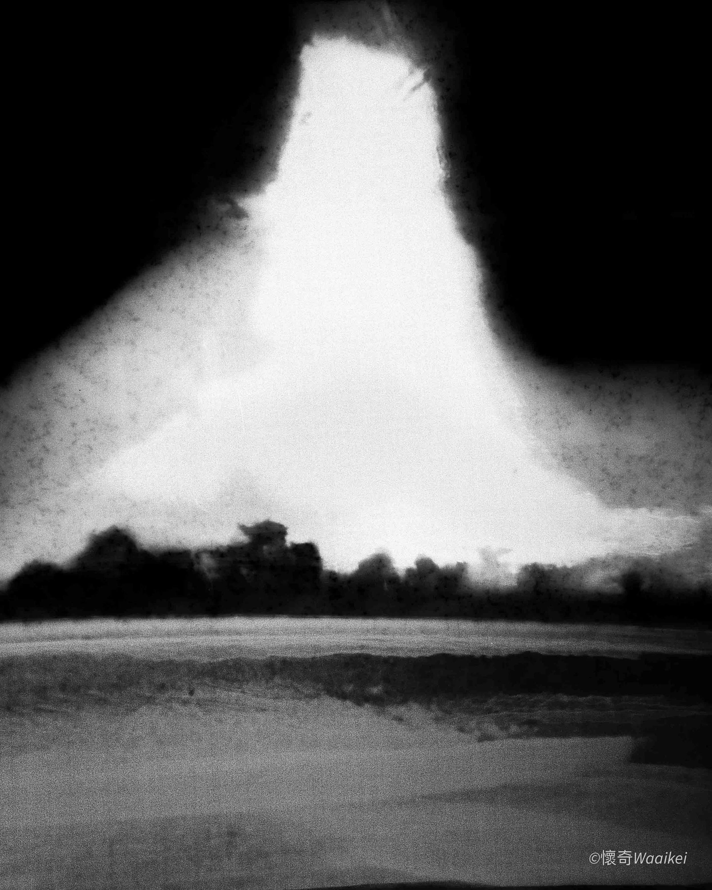 Black and white photo of a large mushroom cloud rising over a landscape with trees and water in the foreground, taken at sunset or sunrise.