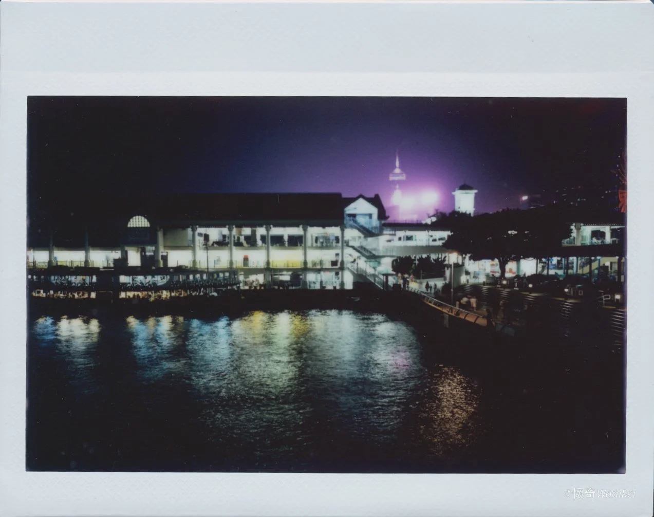 Nighttime view of a waterfront area with lit buildings and a tower in the background, reflecting on the water.