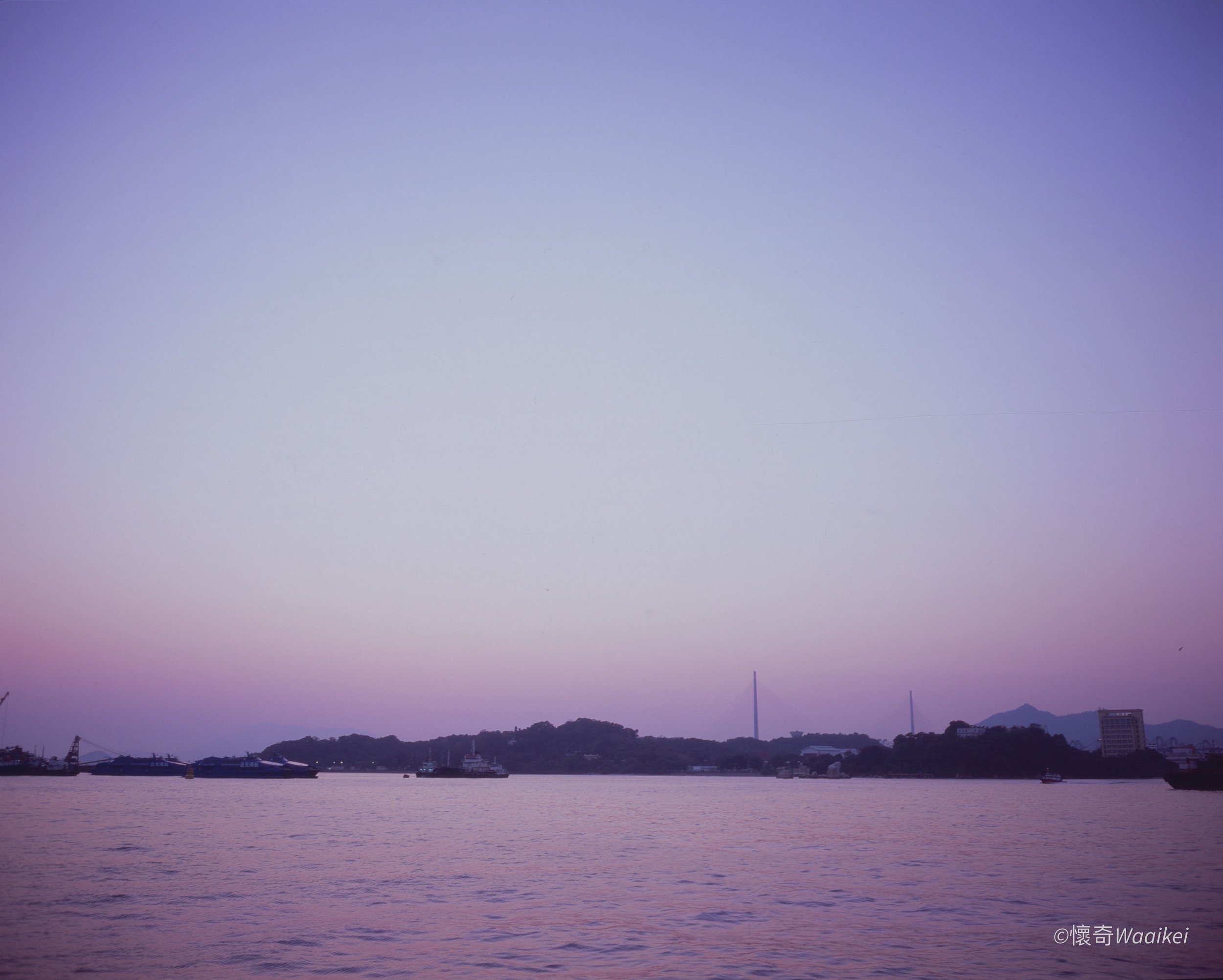 A peaceful water scene at dusk with a city skyline and mountains in the background, purple and pink hues in the sky, and boats on the water.