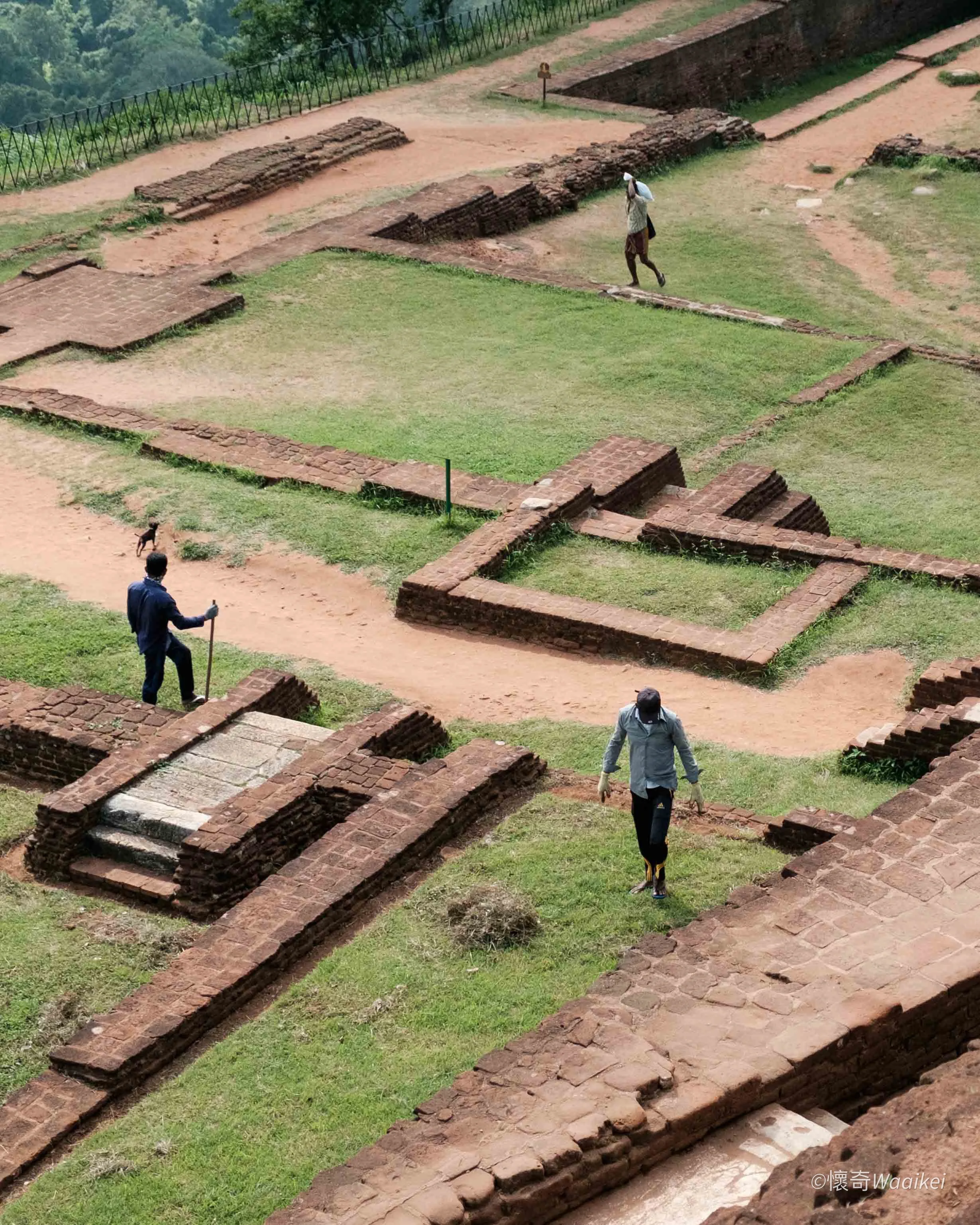 Archaeological site with ancient brick structures and pathways, surrounded by greenery and metal fence, with people exploring the area.