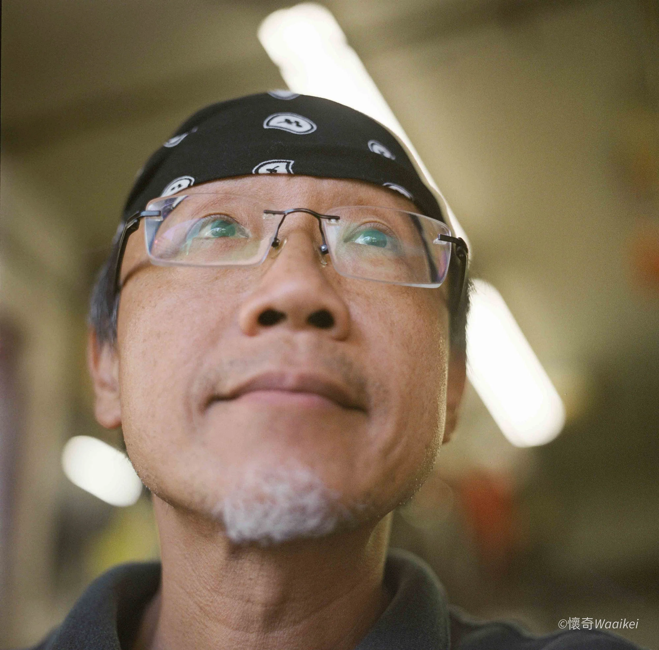 Close-up of a person wearing glasses and a black bandana with white patterns, looking upward with reflections in their glasses, with a blurred background.