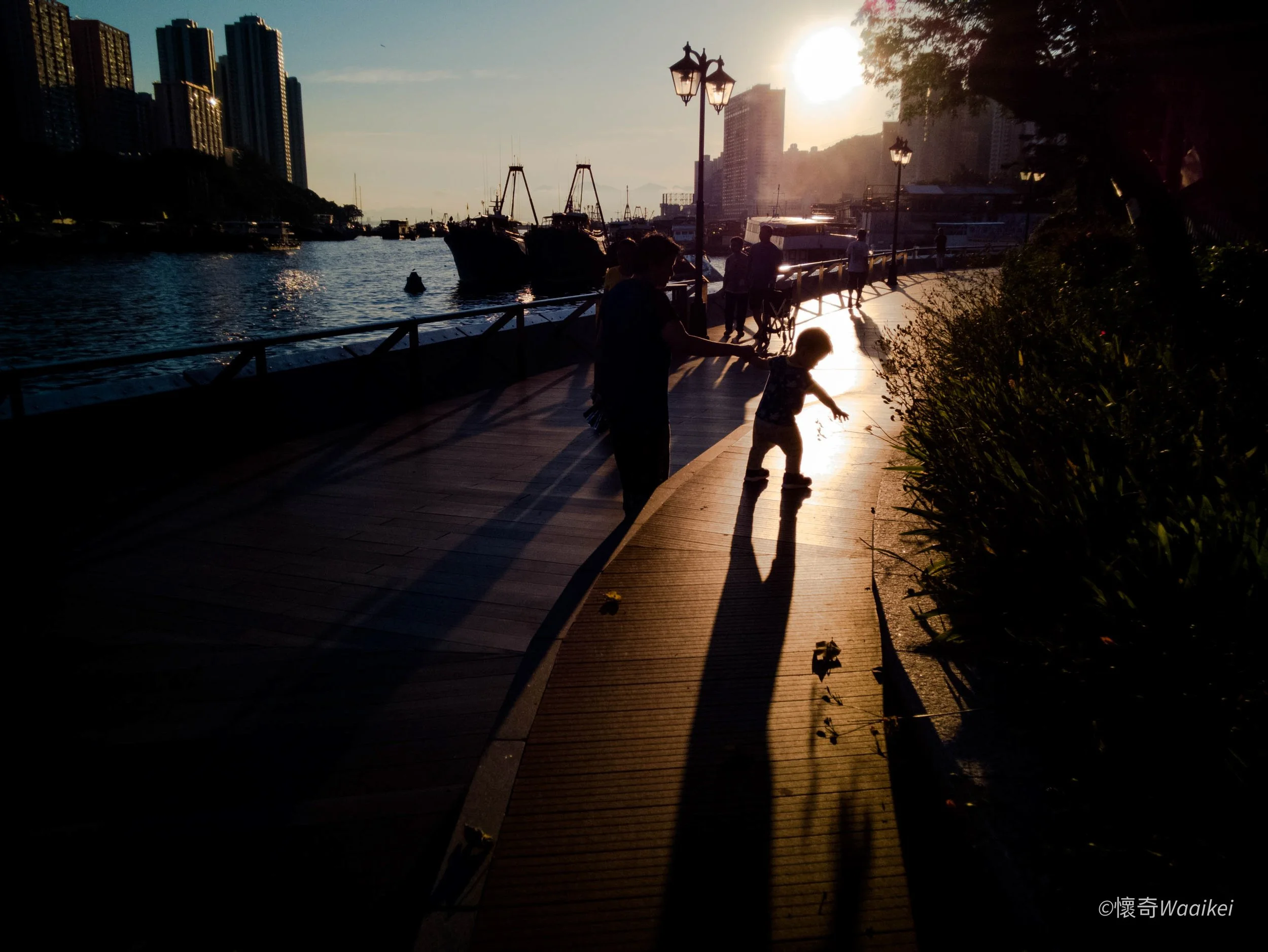 A waterfront scene at sunset with people walking along a wooden pier, boats docked in the water, tall buildings in the background, and trees on the right side.