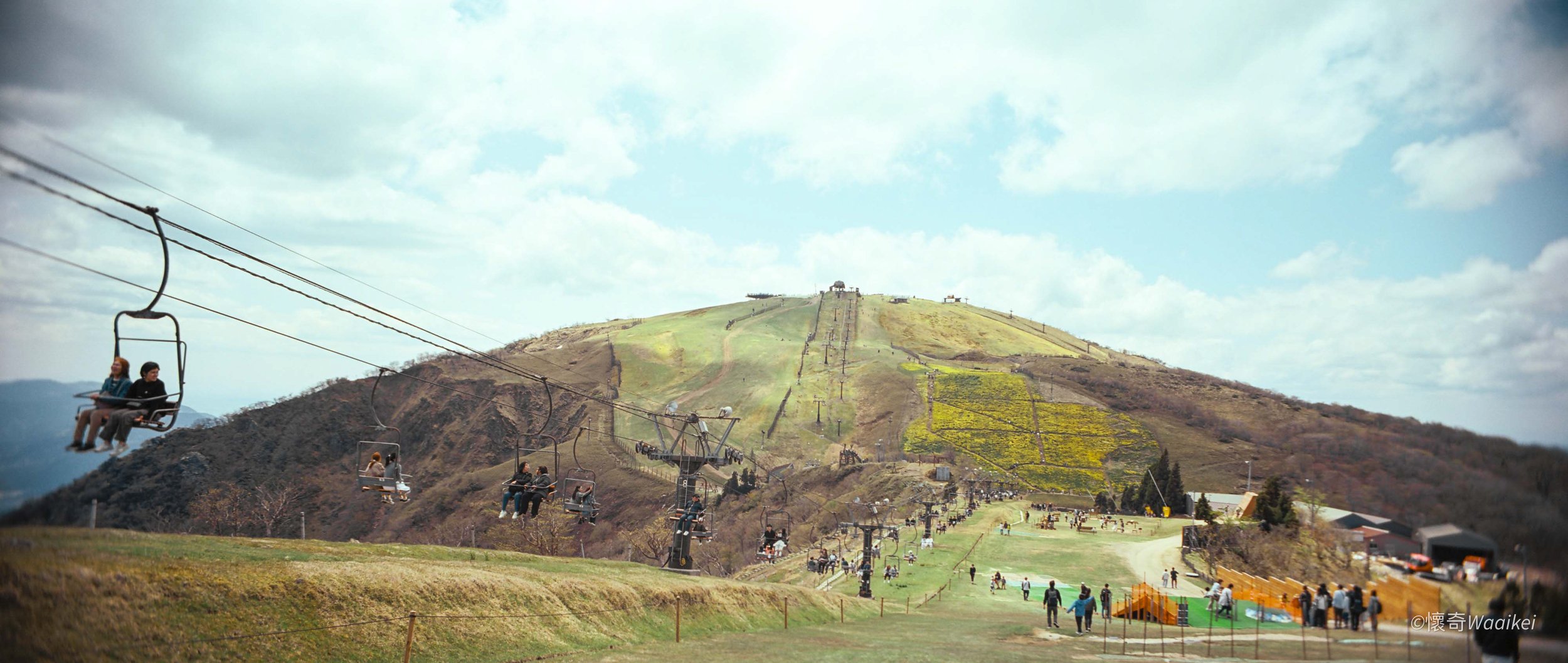 People riding a scenic chairlift over a green hillside with a mountain in the background under partly cloudy sky.
