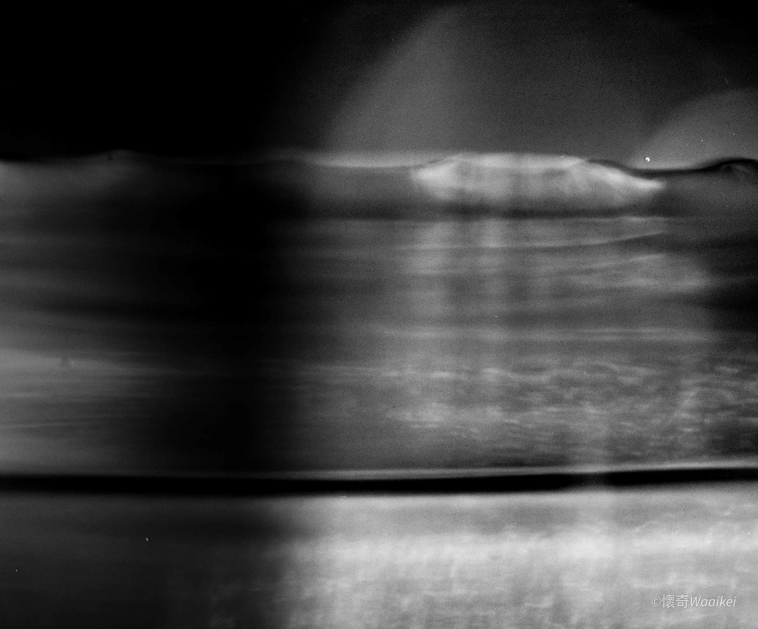 A black and white photo showing ripples on the surface of water at night, with lights shining in the distance across the water.