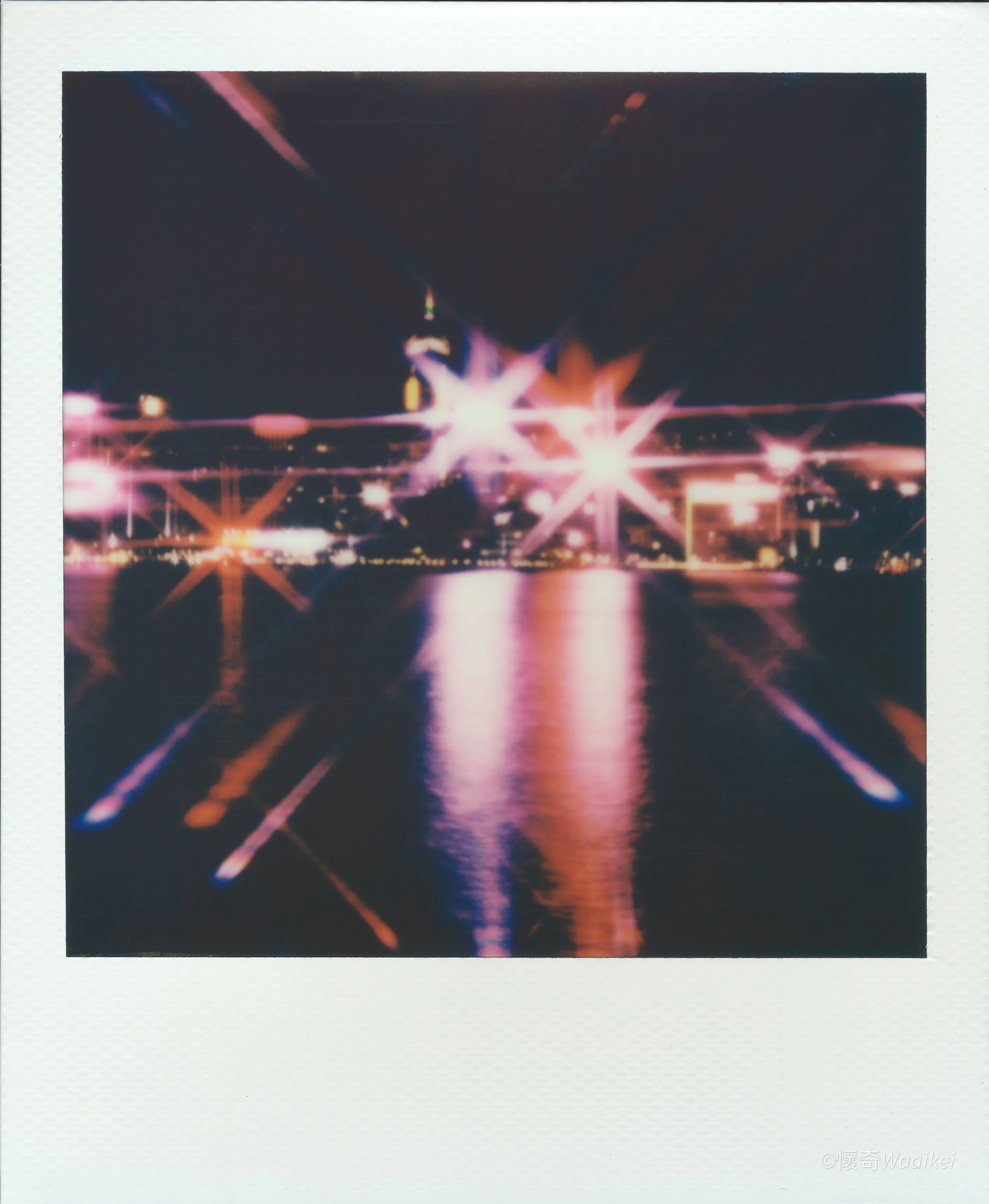 Nighttime cityscape with bright, colorful lights reflected on water, creating starburst effects.