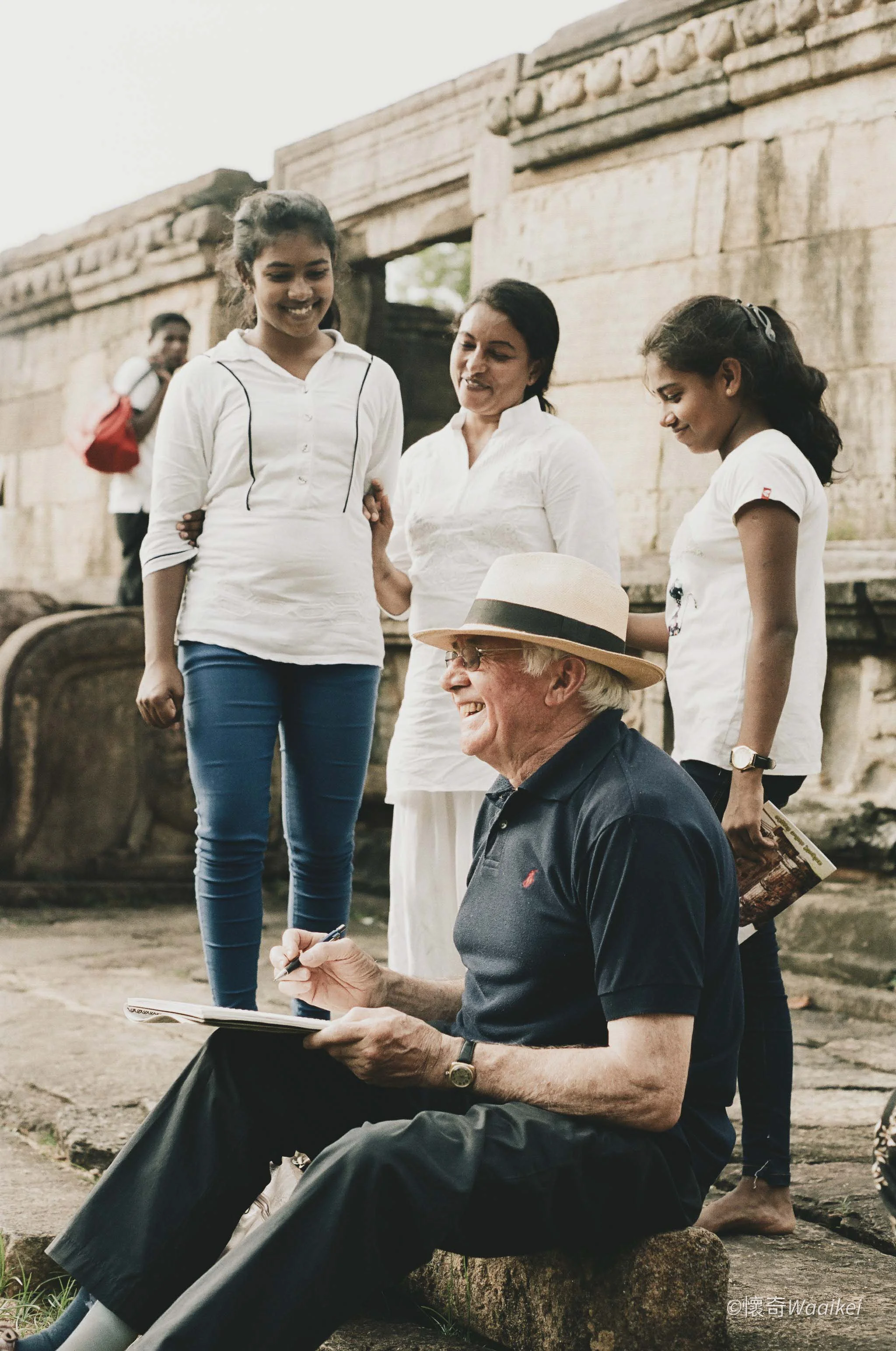 A group of people visiting an ancient archaeological site. An elderly man is seated, signing a book, while three younger women stand nearby, observing and smiling. The background shows stone ruins and a person with a backpack is visible in the distan