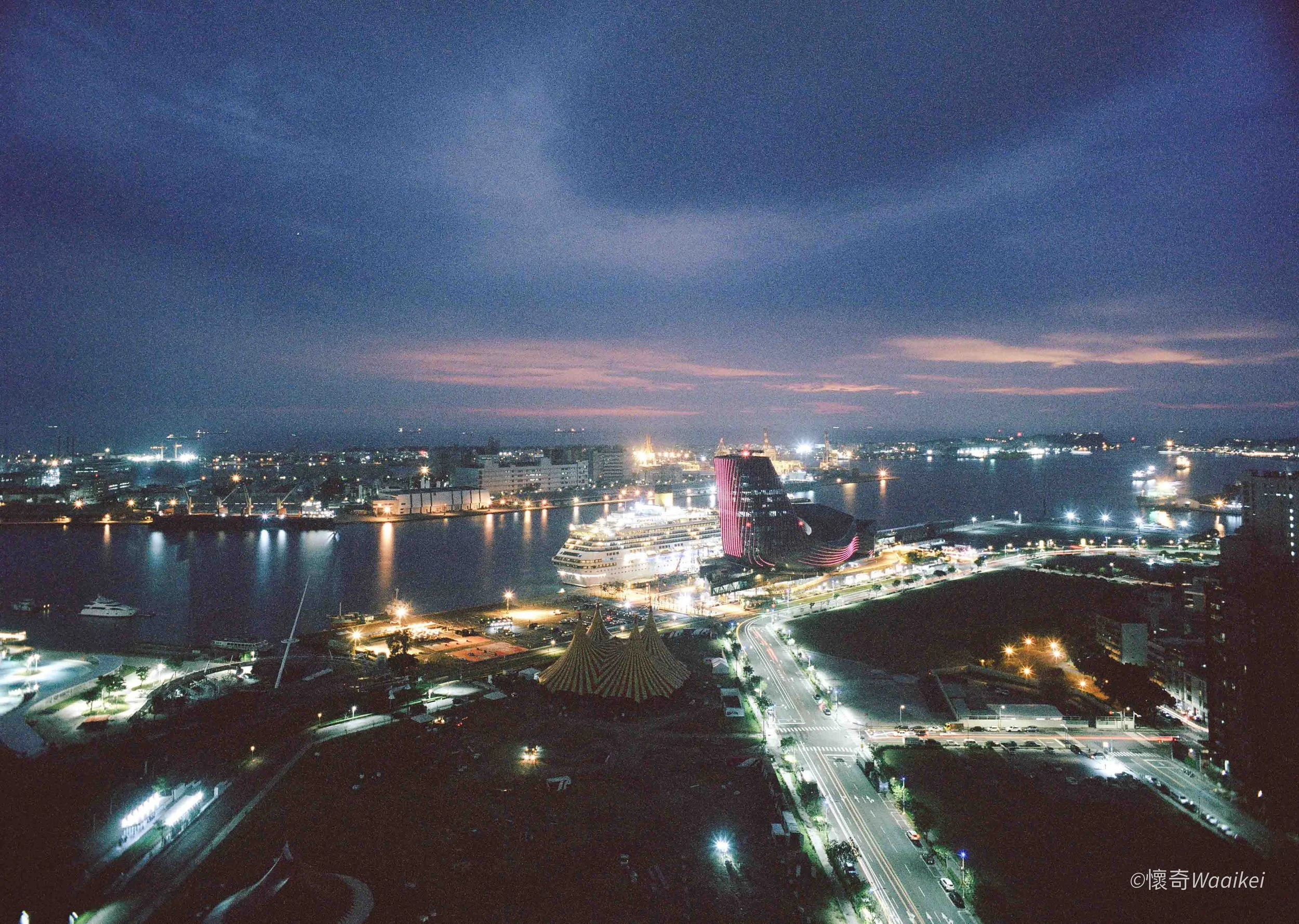 Nighttime cityscape view of a harbor with illuminated buildings, boats, and a cruise ship, under a dark sky with clouds.