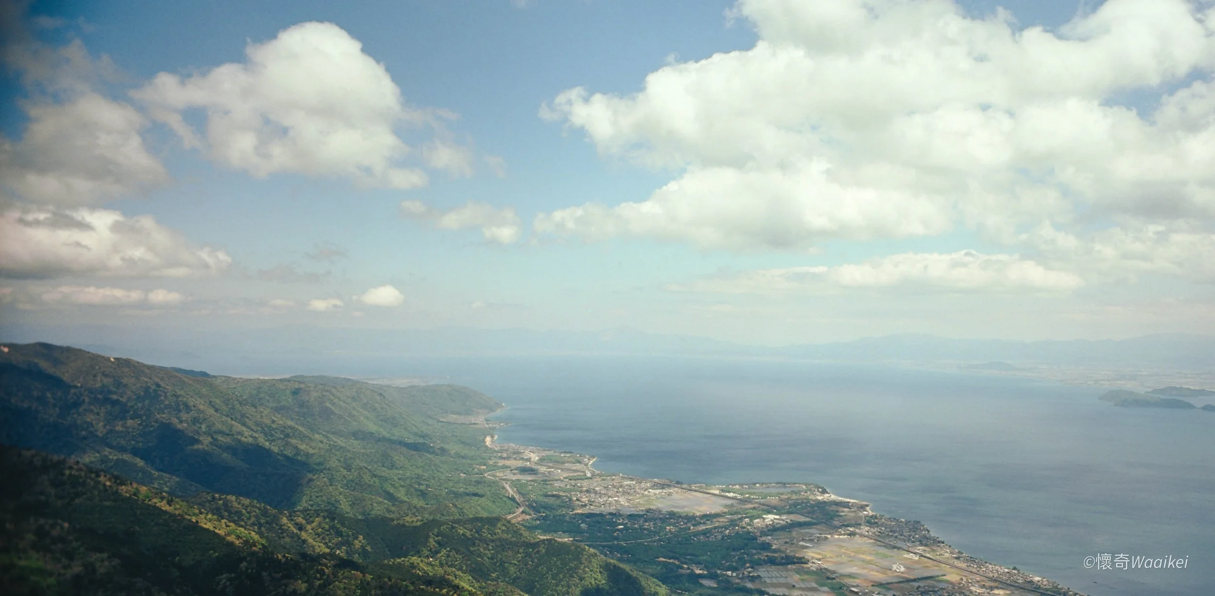 Aerial view of green mountains descending to coastal area with water, clouds in sky