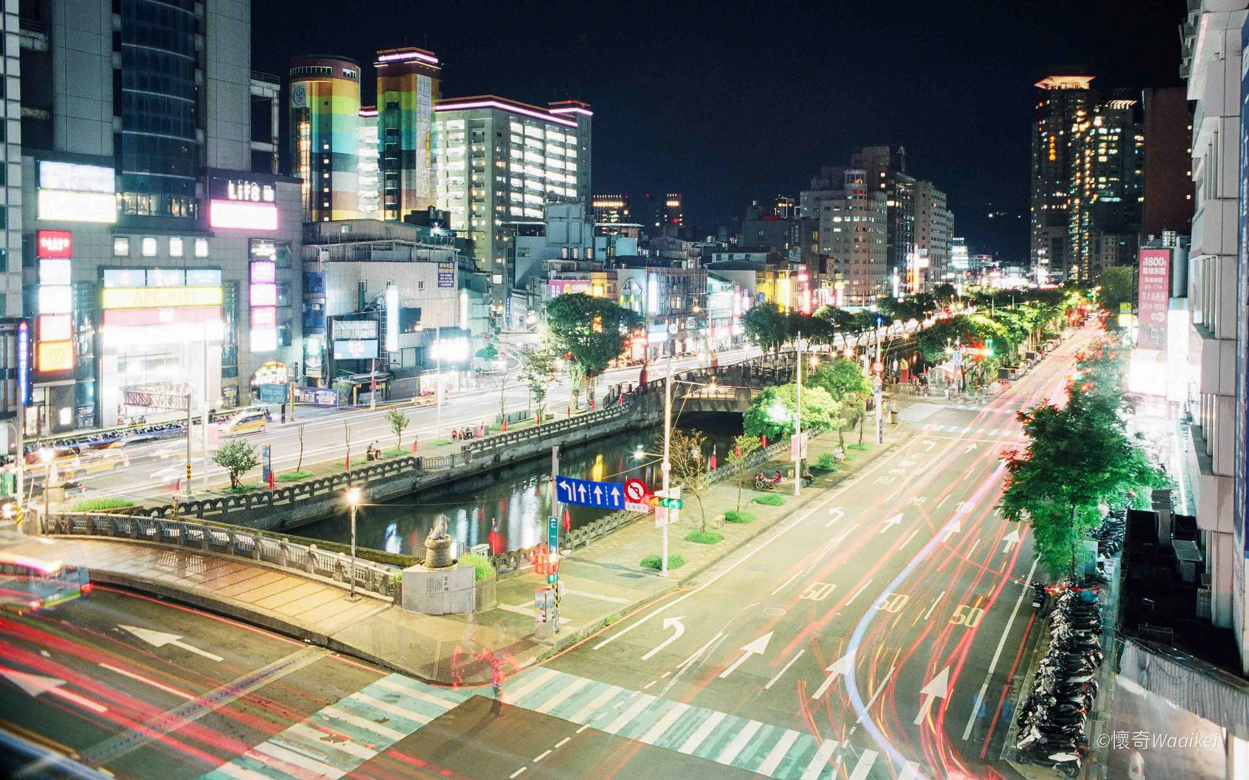 Nighttime city street scene with illuminated buildings, moving vehicles with light trails, and crosswalks.
