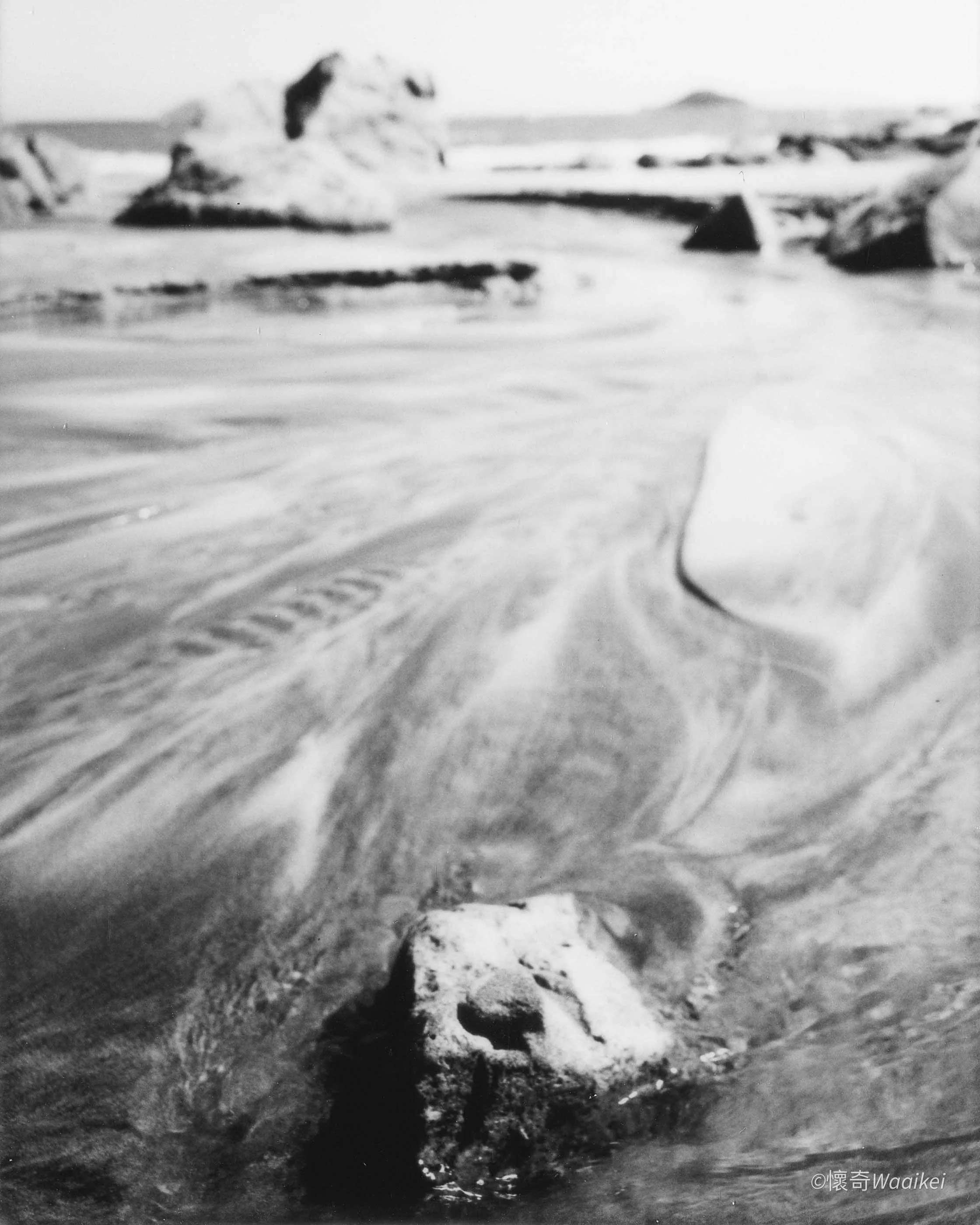 Black and white photo of a rugged beach scene with rocks and wet sand, taken from a low angle, featuring a sizable rock formation in the background, and a small sign or object partially buried in the sand in the foreground, with a textured shoreline 
