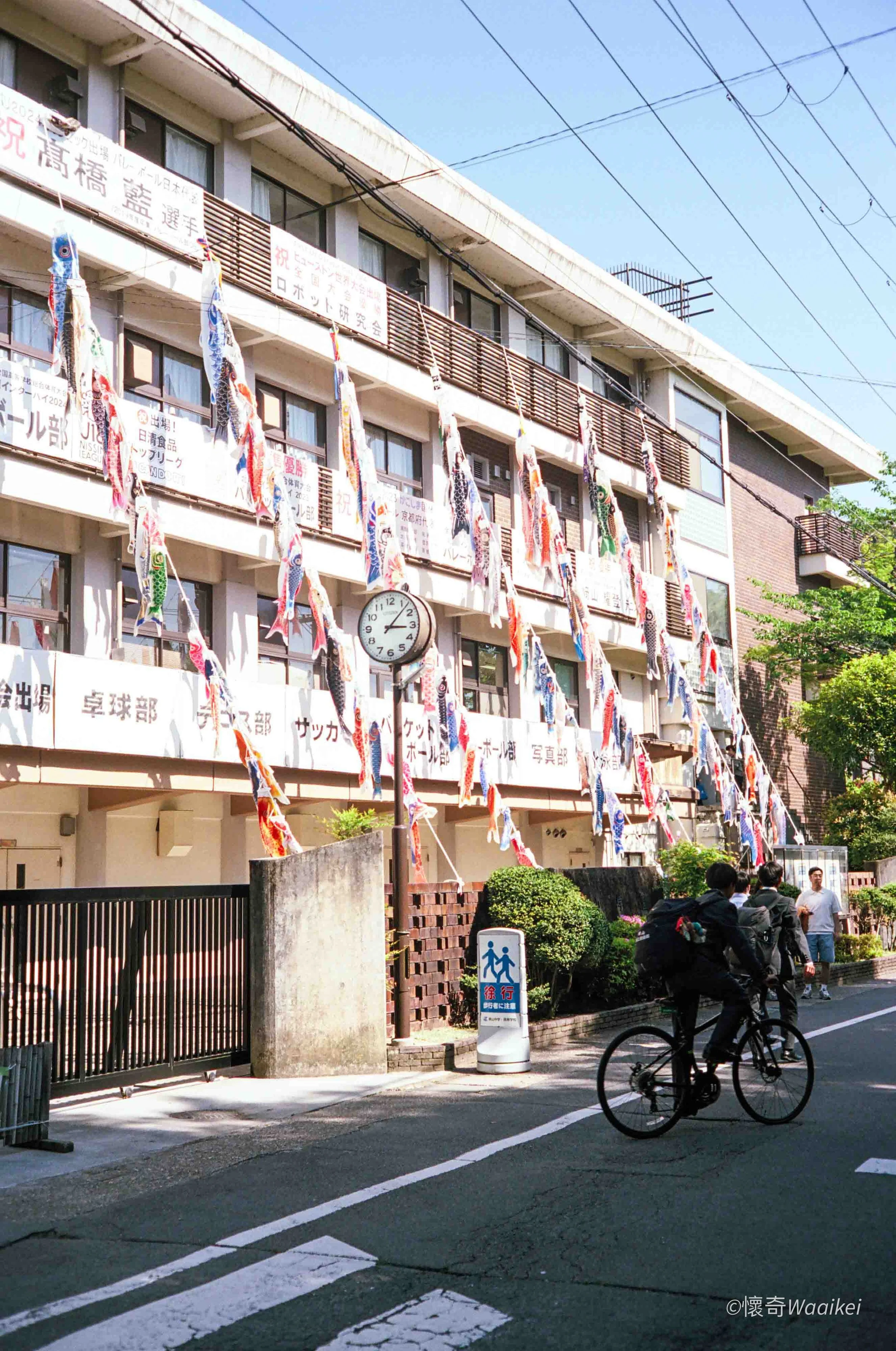 A multi-story building decorated with colorful carp-shaped streamers and banners, with pedestrians and cyclists on the street in front.
