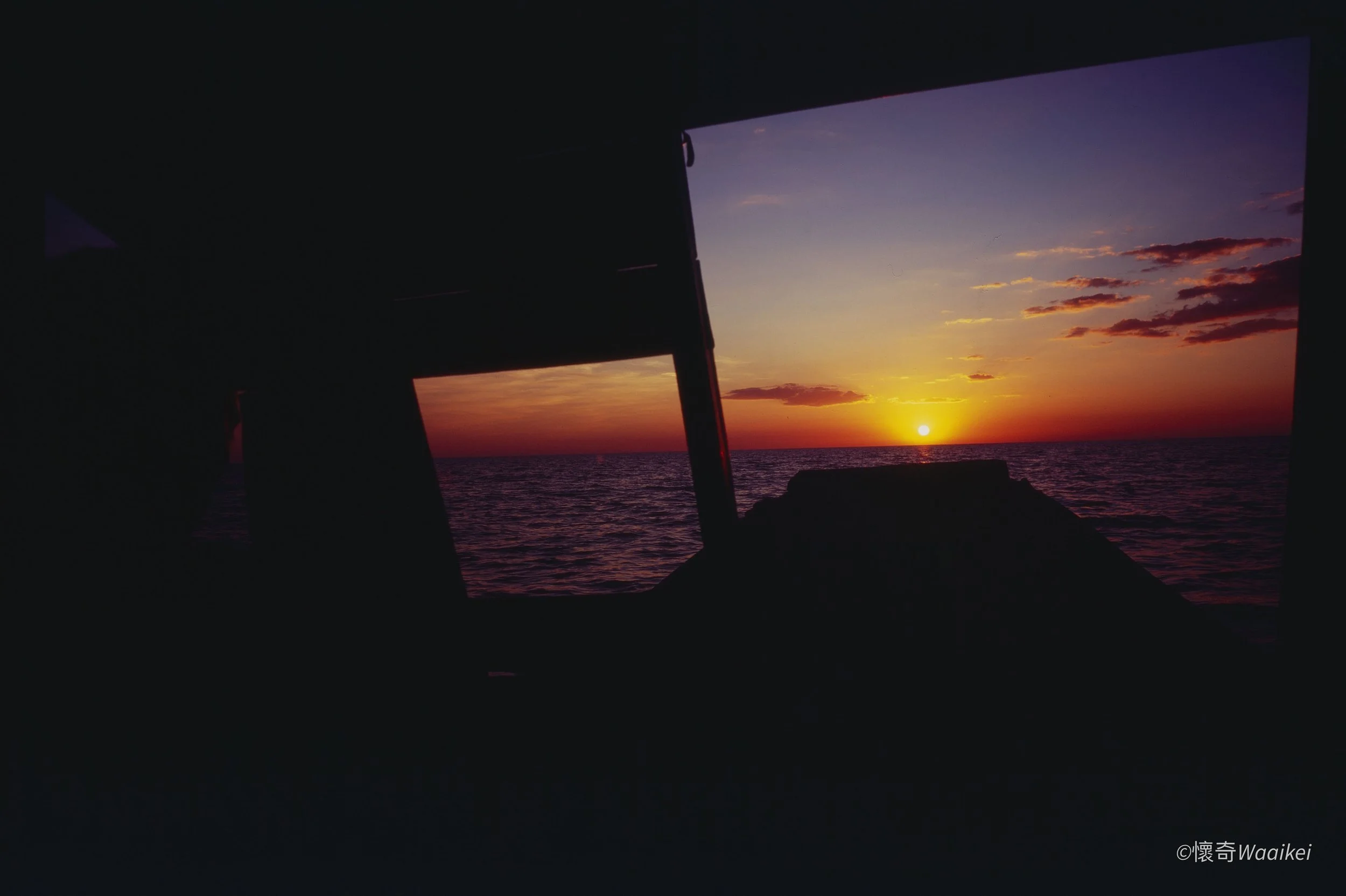 A sunset over the ocean viewed through the windows of a dark room or structure, with the sun near the horizon and colorful clouds in the sky.