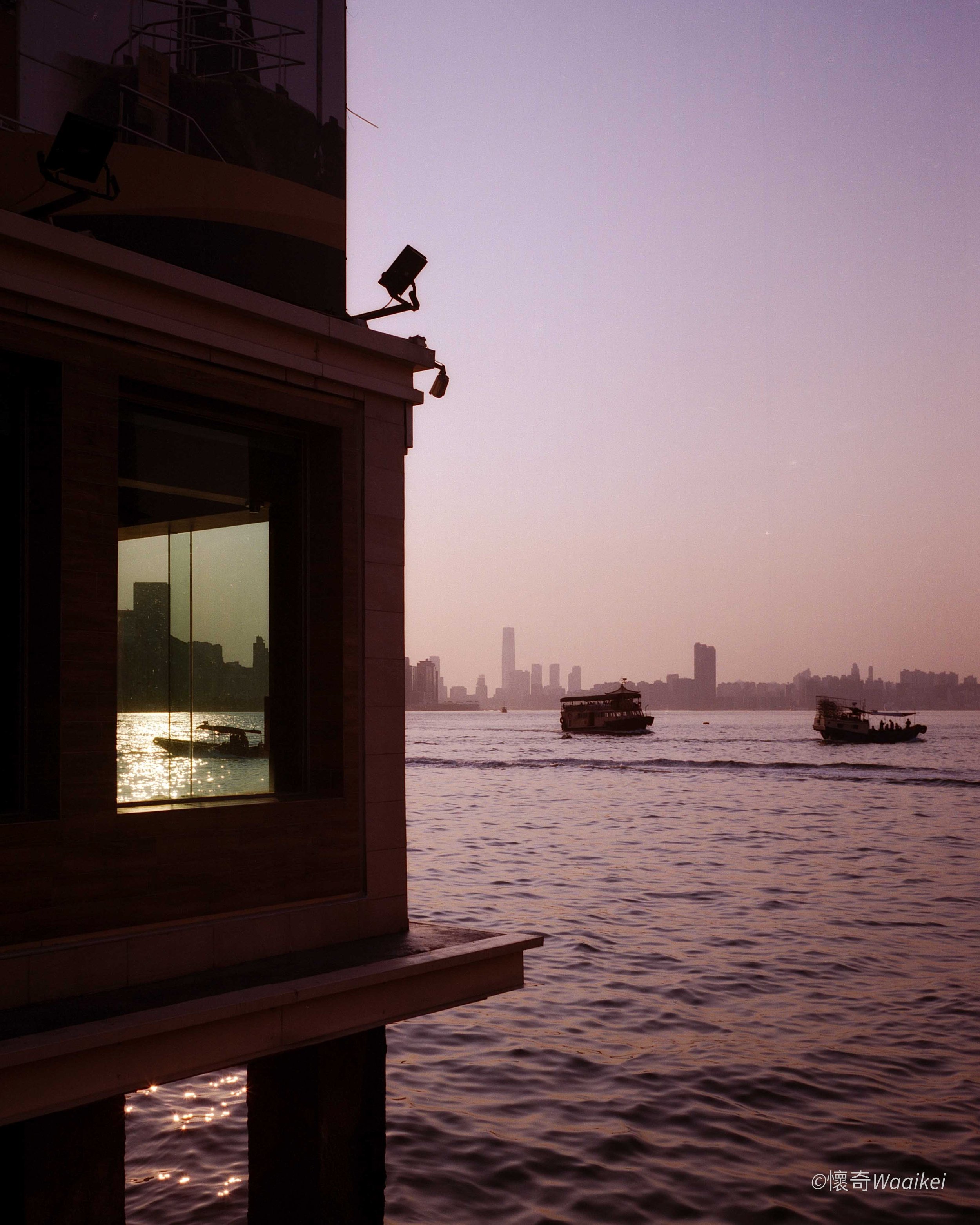 Silhouette of a building with large window and outdoor lights, overlooking a body of water with boats, and a city skyline in the sunset or sunrise.