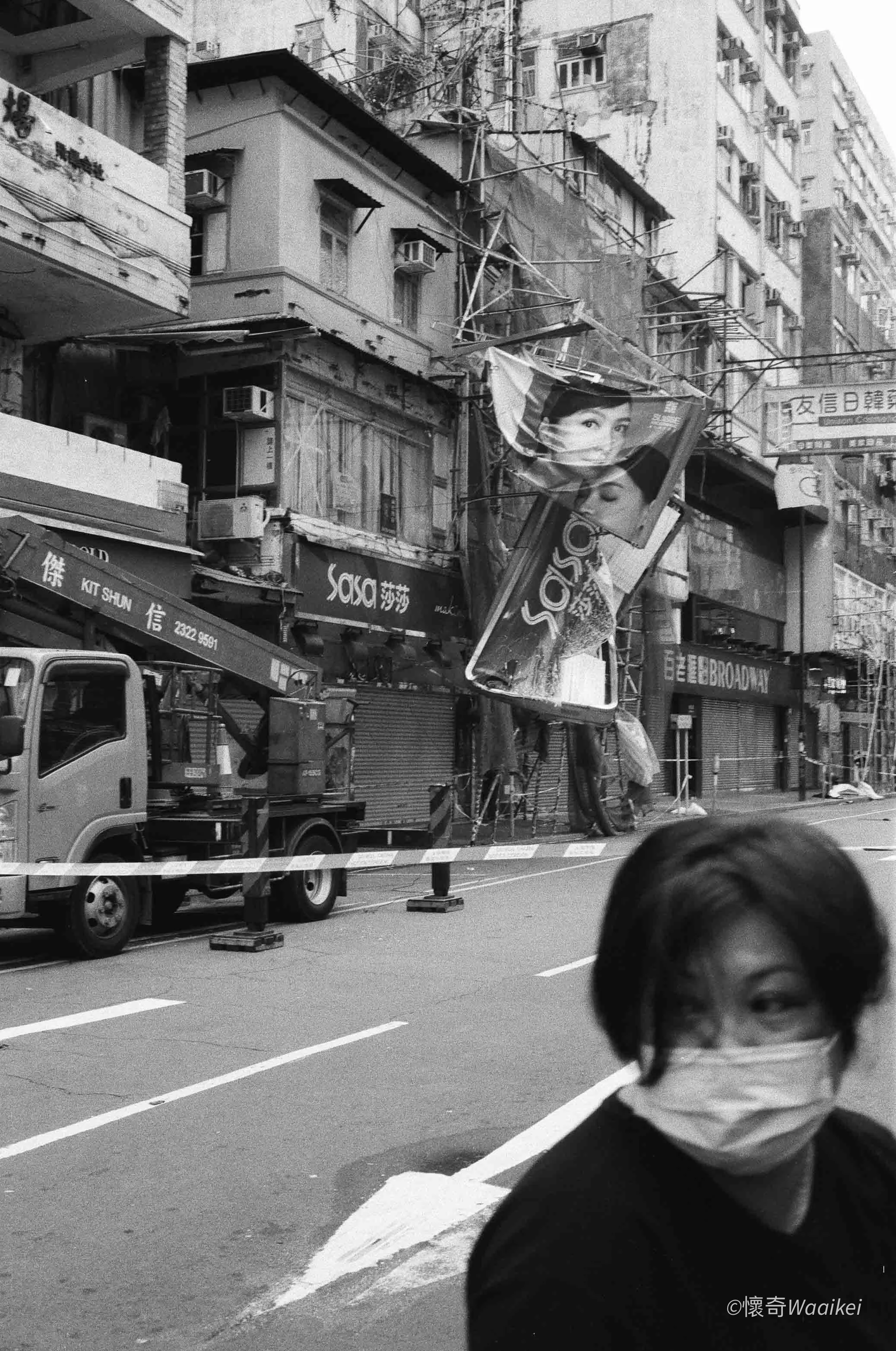 Street scene in an urban area with storefronts and buildings, some under construction or renovation, with a person wearing a face mask in the foreground.