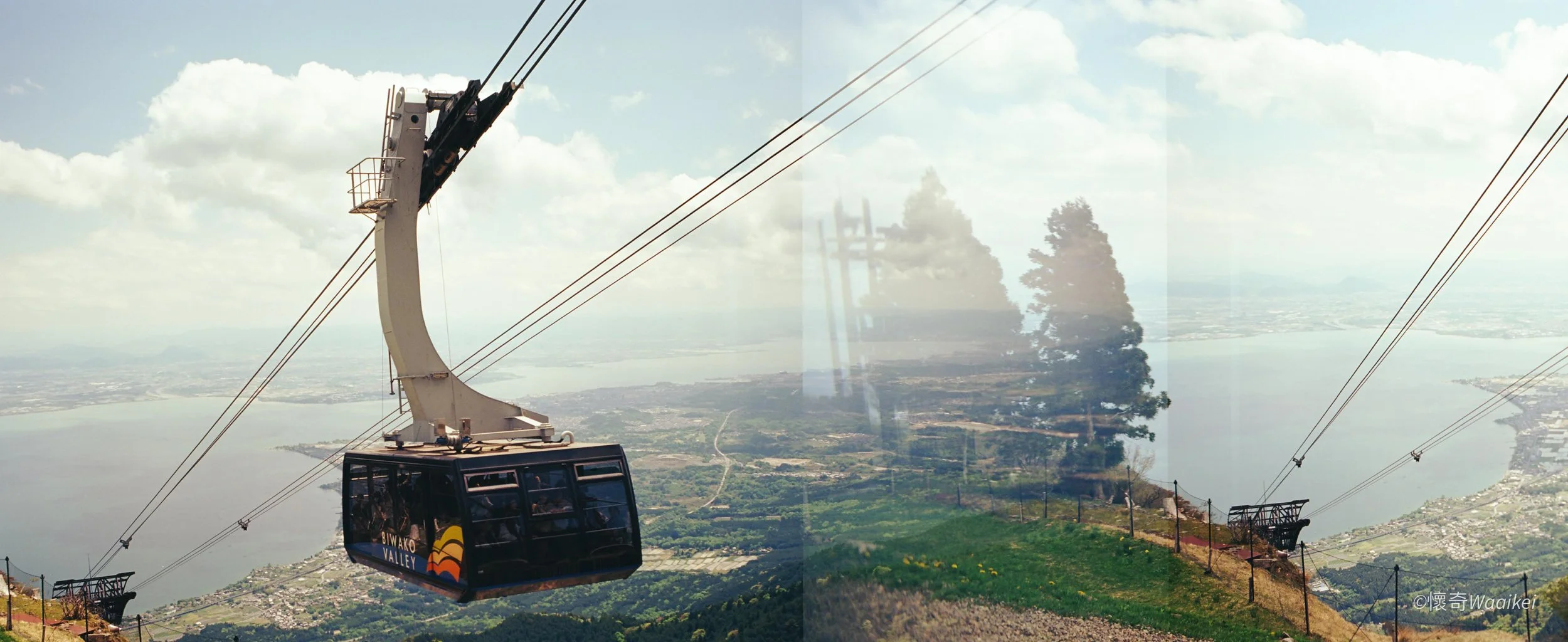 A cable car labeled 'Biwako Valley' ascending over green hills with a view of a river and distant cityscape under a partly cloudy sky.