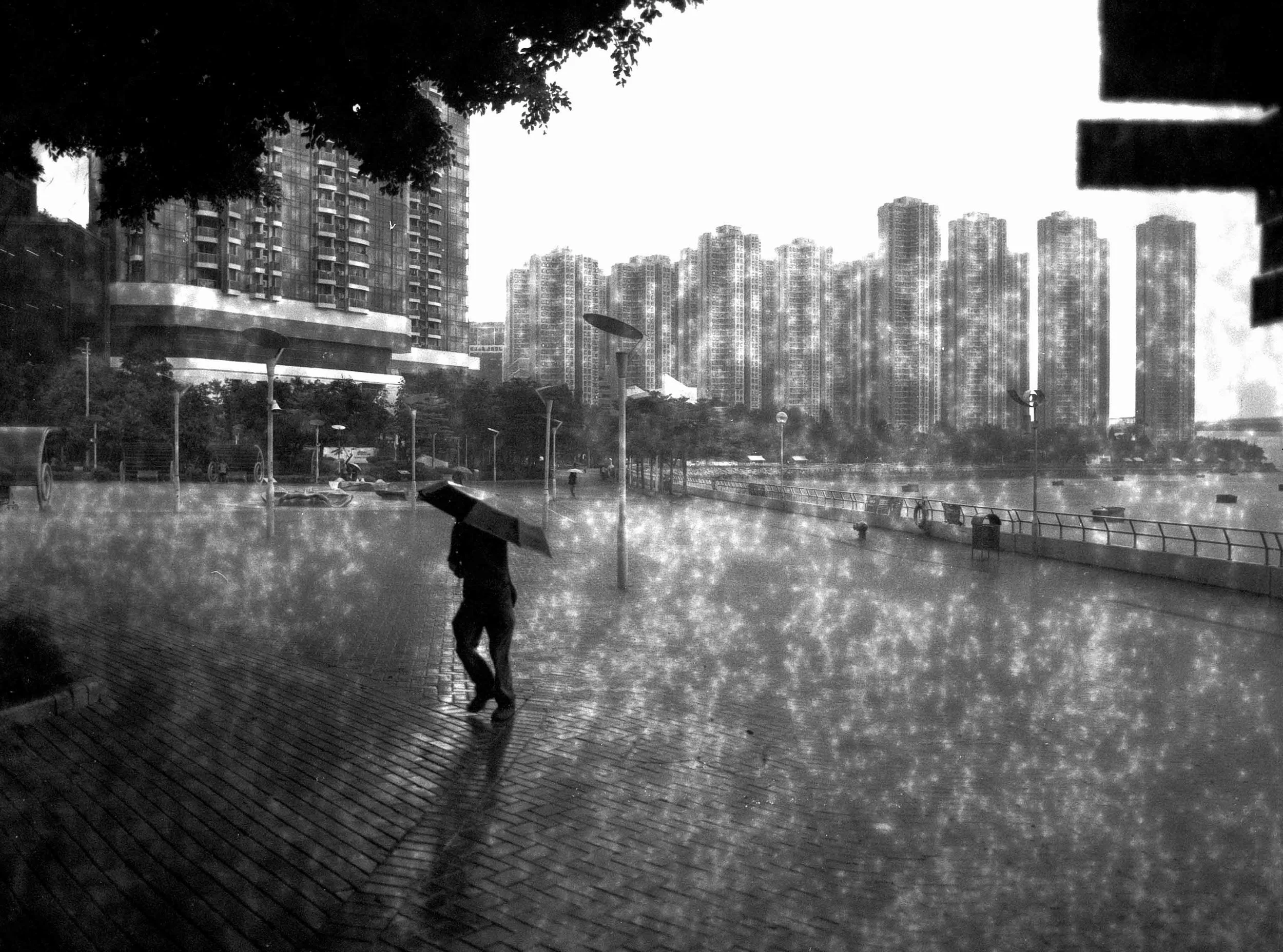 A person holding an umbrella walking on a wet sidewalk during rain at dusk, with high-rise buildings in the background.