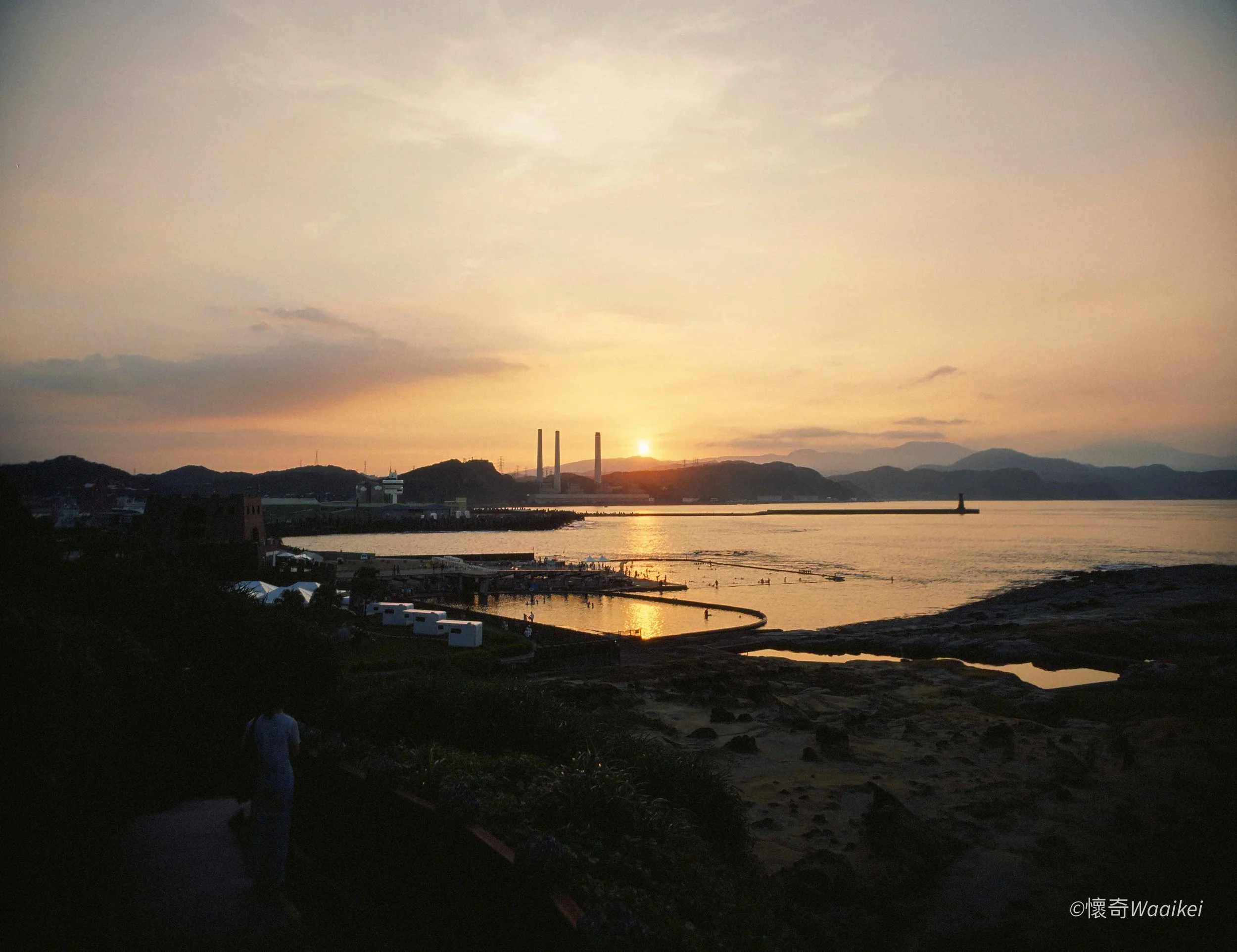 Sunset over a calm body of water with mountains, industrial chimneys, and people on the shore.