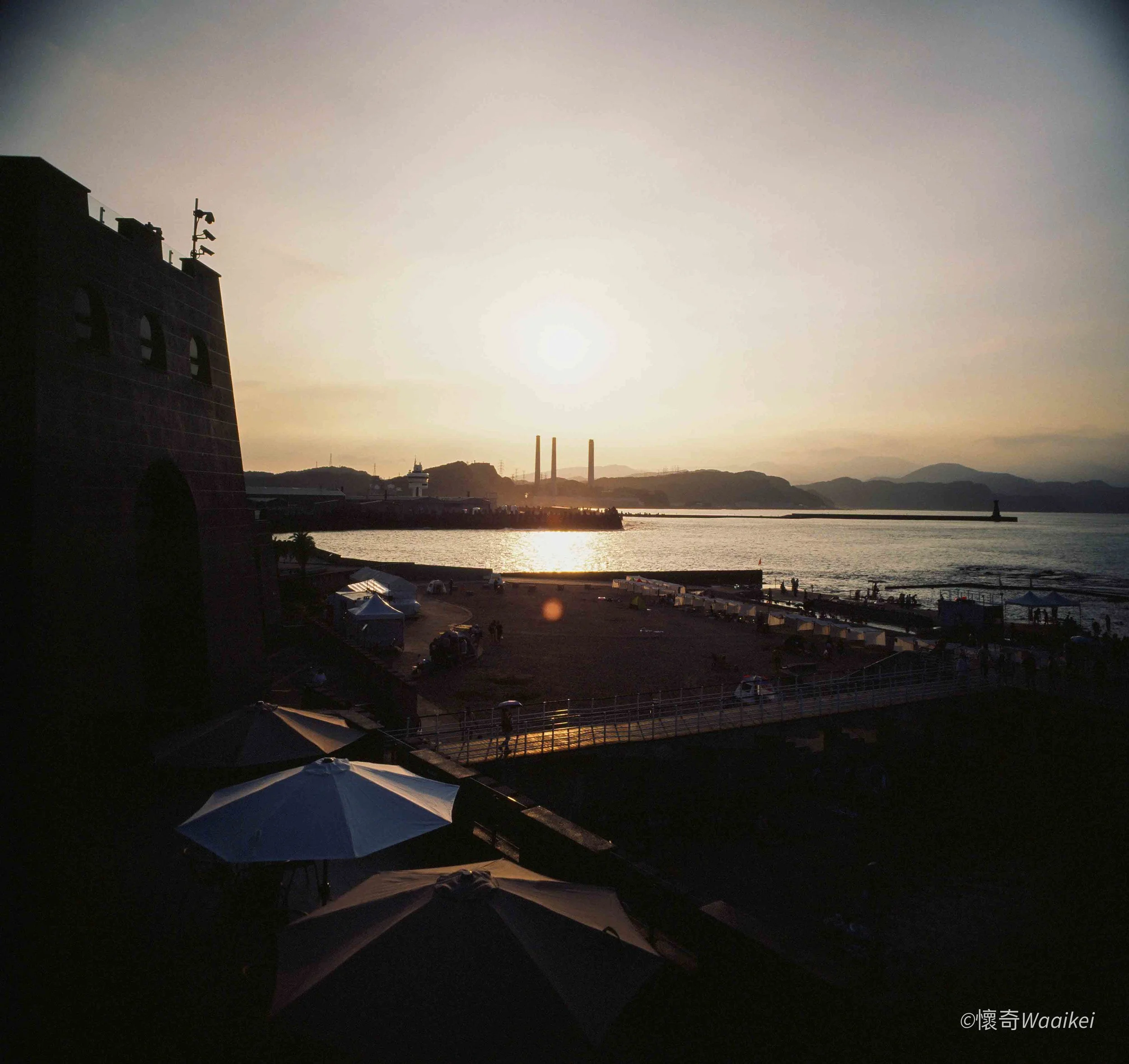 Sunset over a harbor with boats, a building with antennas, and mountains in the background.