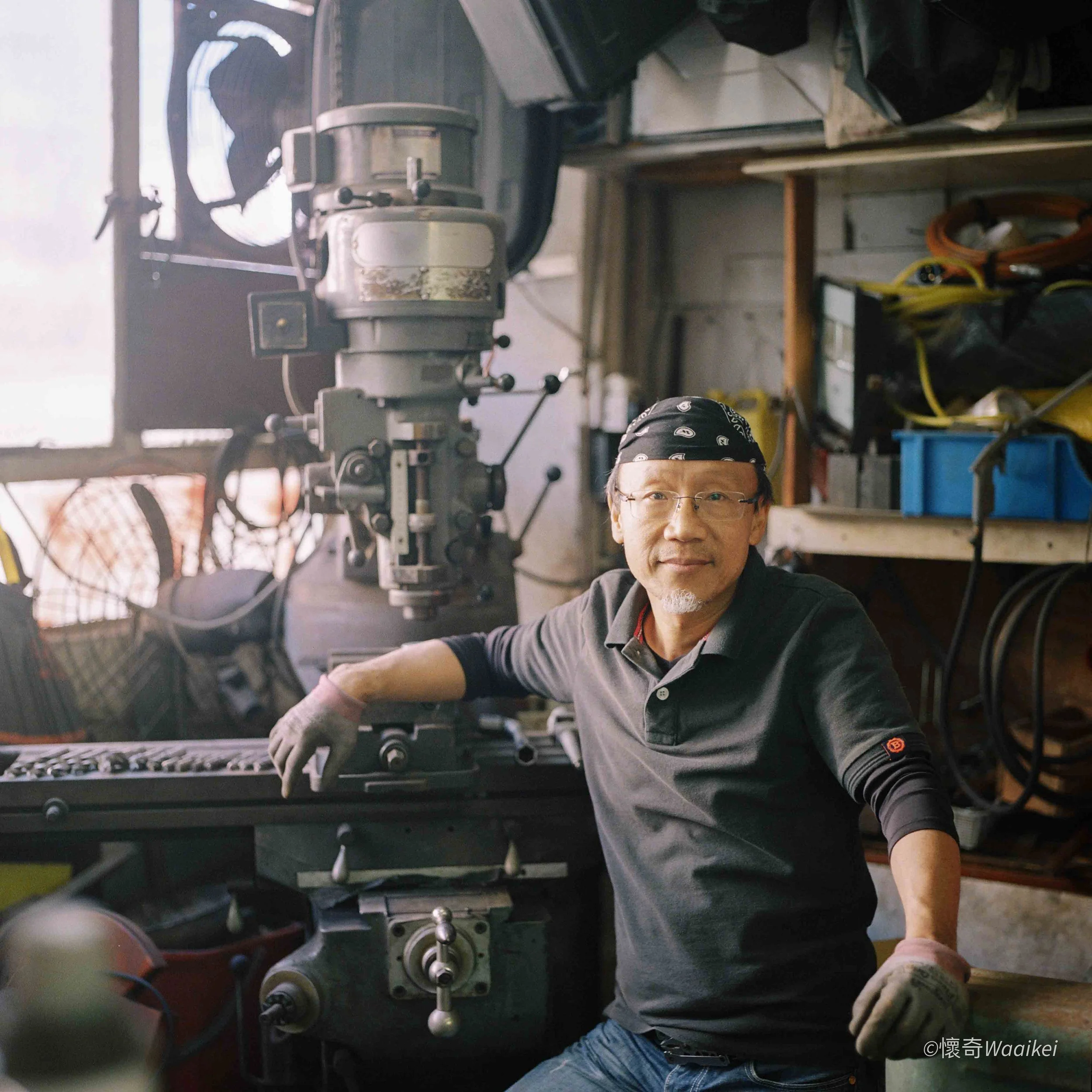 A man wearing glasses, a bandana, and a gray shirt working in a workshop with a milling machine. Shelves with tools and wires are visible in the background.