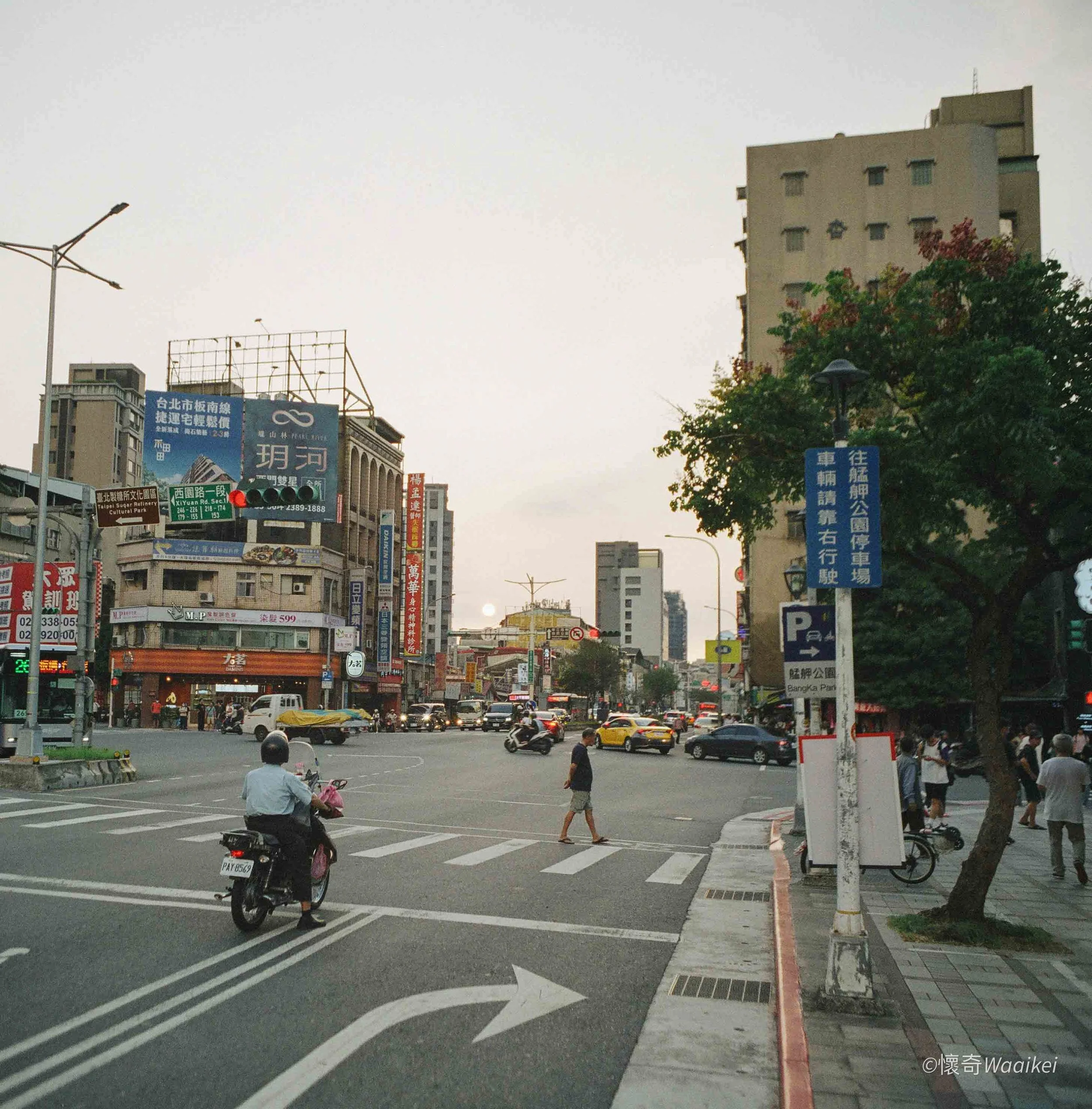 City street scene during the daytime with cars, scooters, and pedestrians. There are tall buildings, street signs, and trees lining the sidewalk. The sky is clear and the sun is setting or rising in the background.