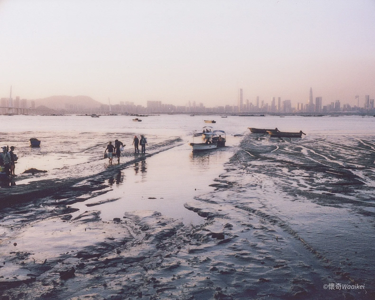 People walking through shallow water with boats in the background and a city skyline in the distance.