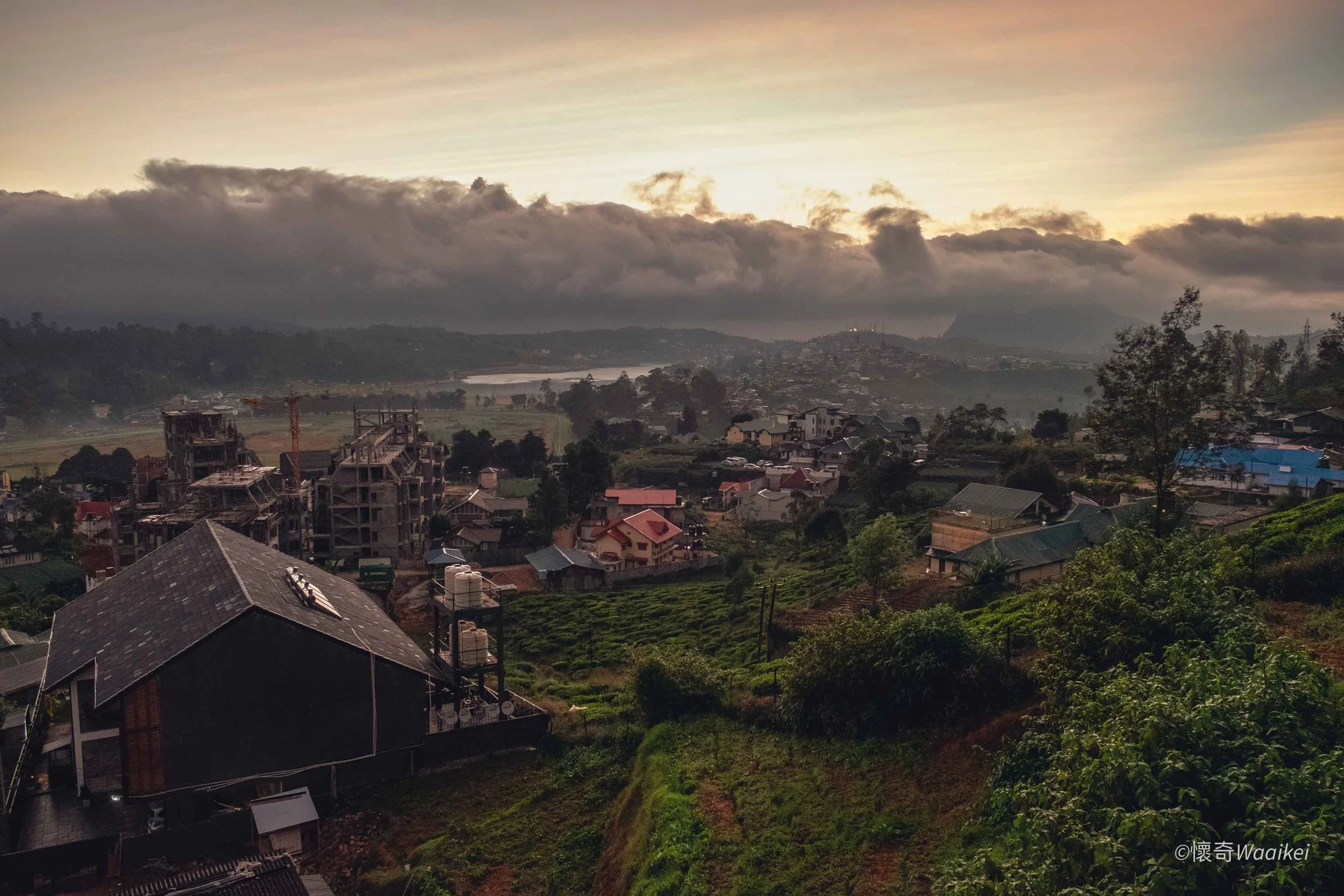 Scenic view of a small town or village nestled in green hills with houses under cloudy sky at sunset or sunrise.