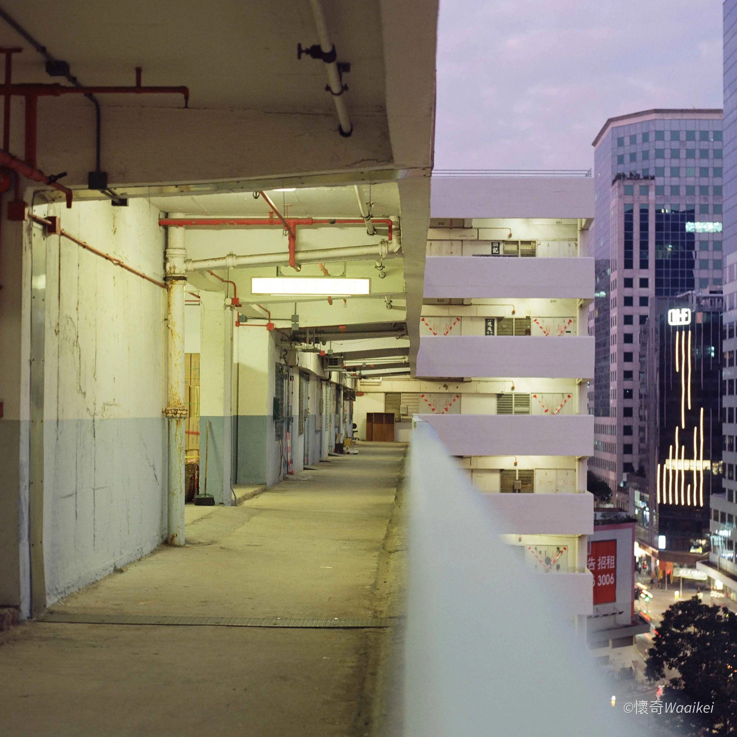 An empty parking garage with exposed pipes and concrete columns, overlooking a cityscape with tall buildings in the background during early evening or dusk.