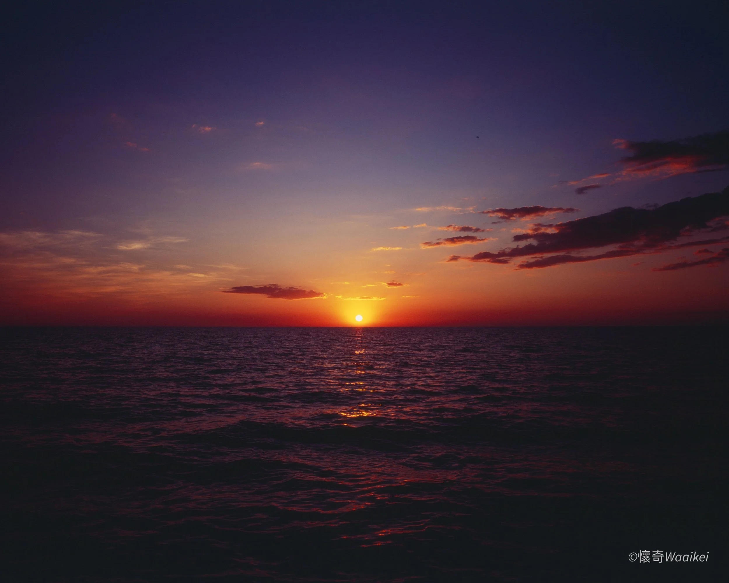 Sun setting over the ocean with a partly cloudy sky, orange and pink hues near the horizon, and dark clouds.