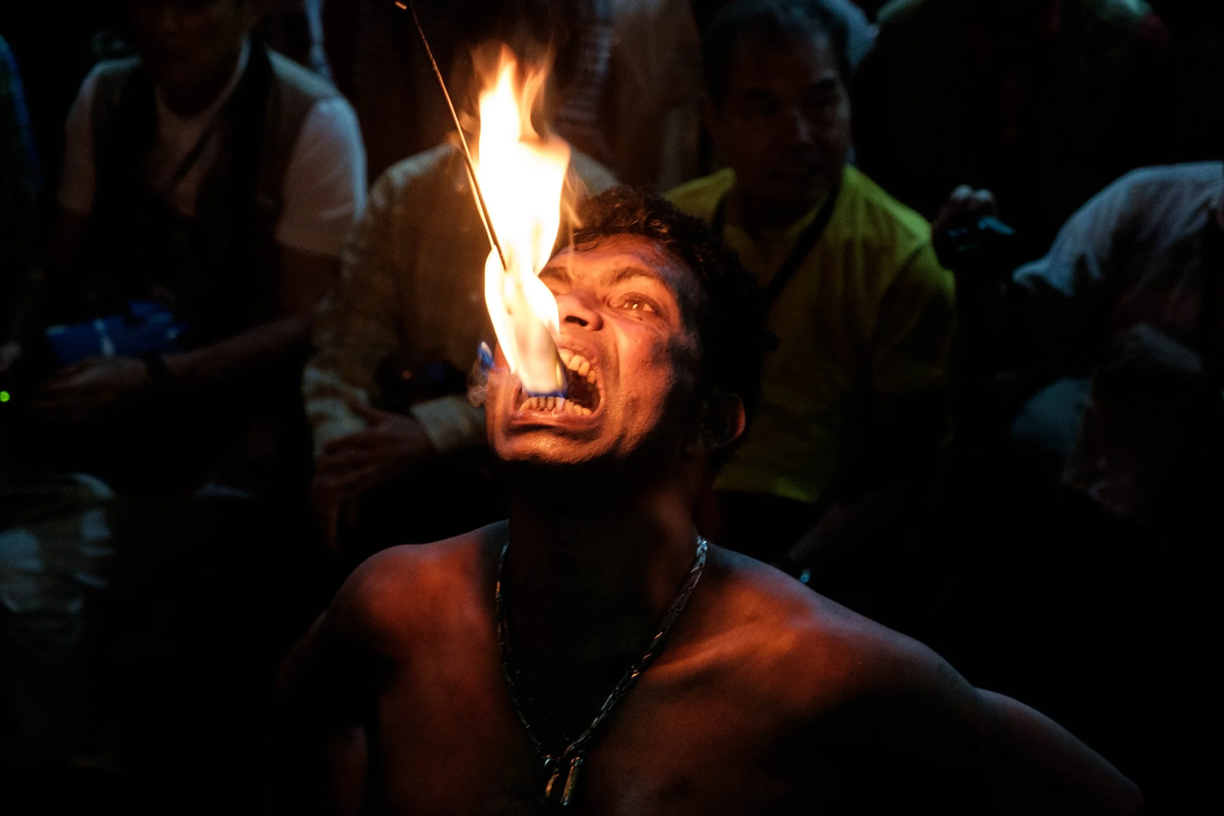 A man with dark curly hair and a chain necklace is performing a fire-eating act, with a flame in his mouth. There are people around him watching in a dark setting.