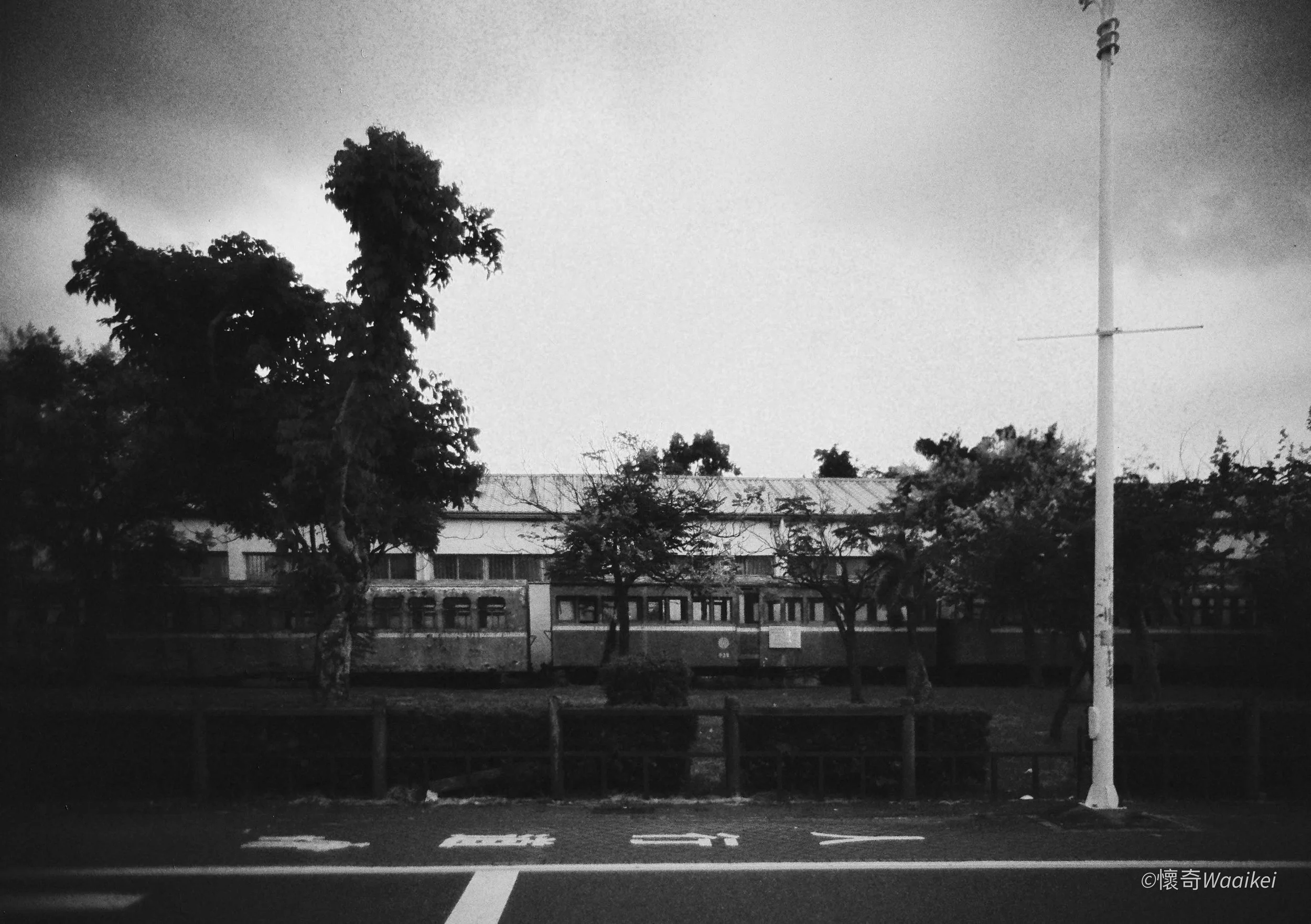 A black-and-white photo of a parking lot with trees, a building in the background, and a tall streetlight post on the right, under a cloudy sky.