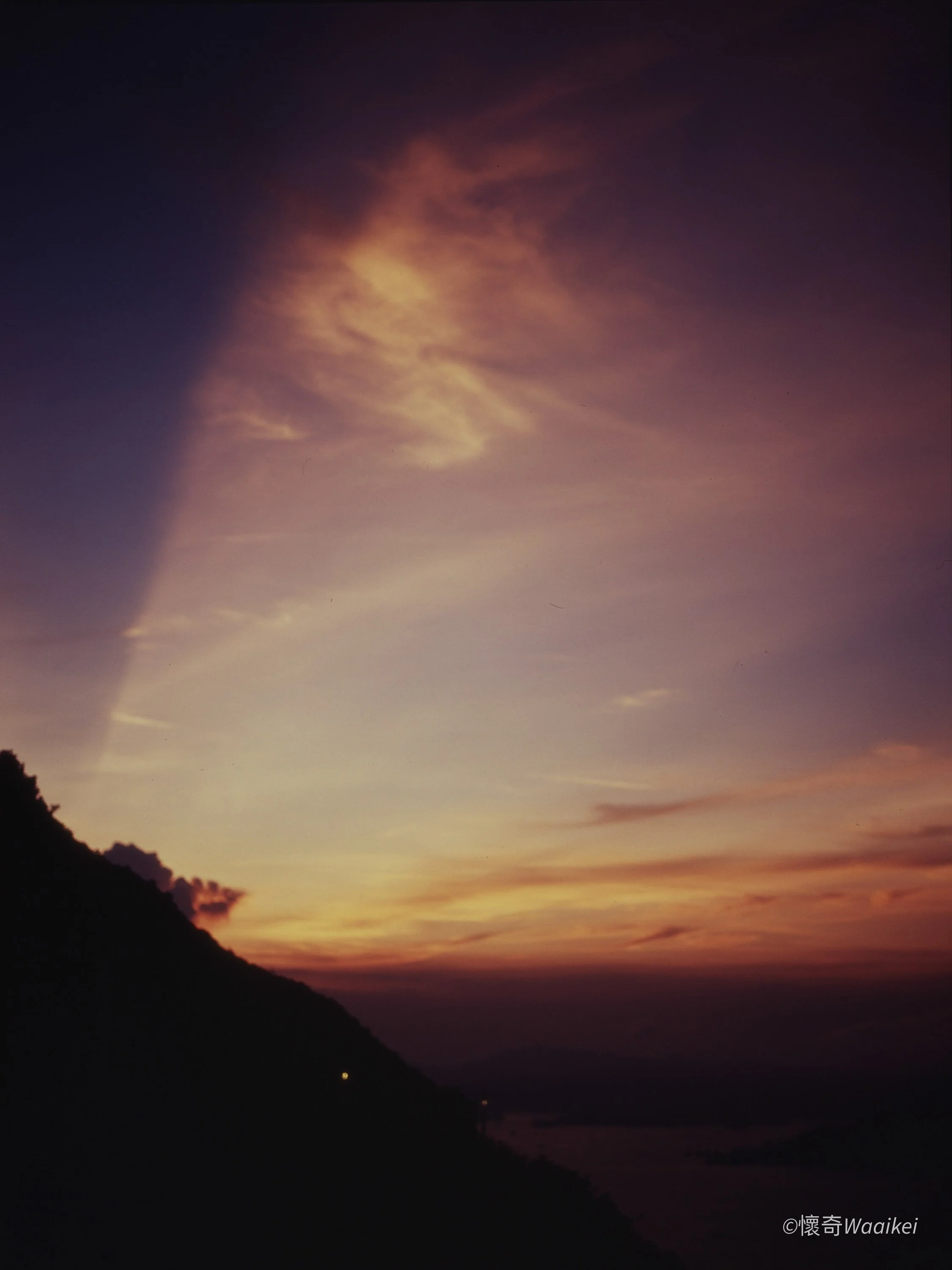 Sunset with a colorful sky blending shades of purple, pink, and orange over a mountainous coastline, with silhouettes of hills and trees in the foreground.