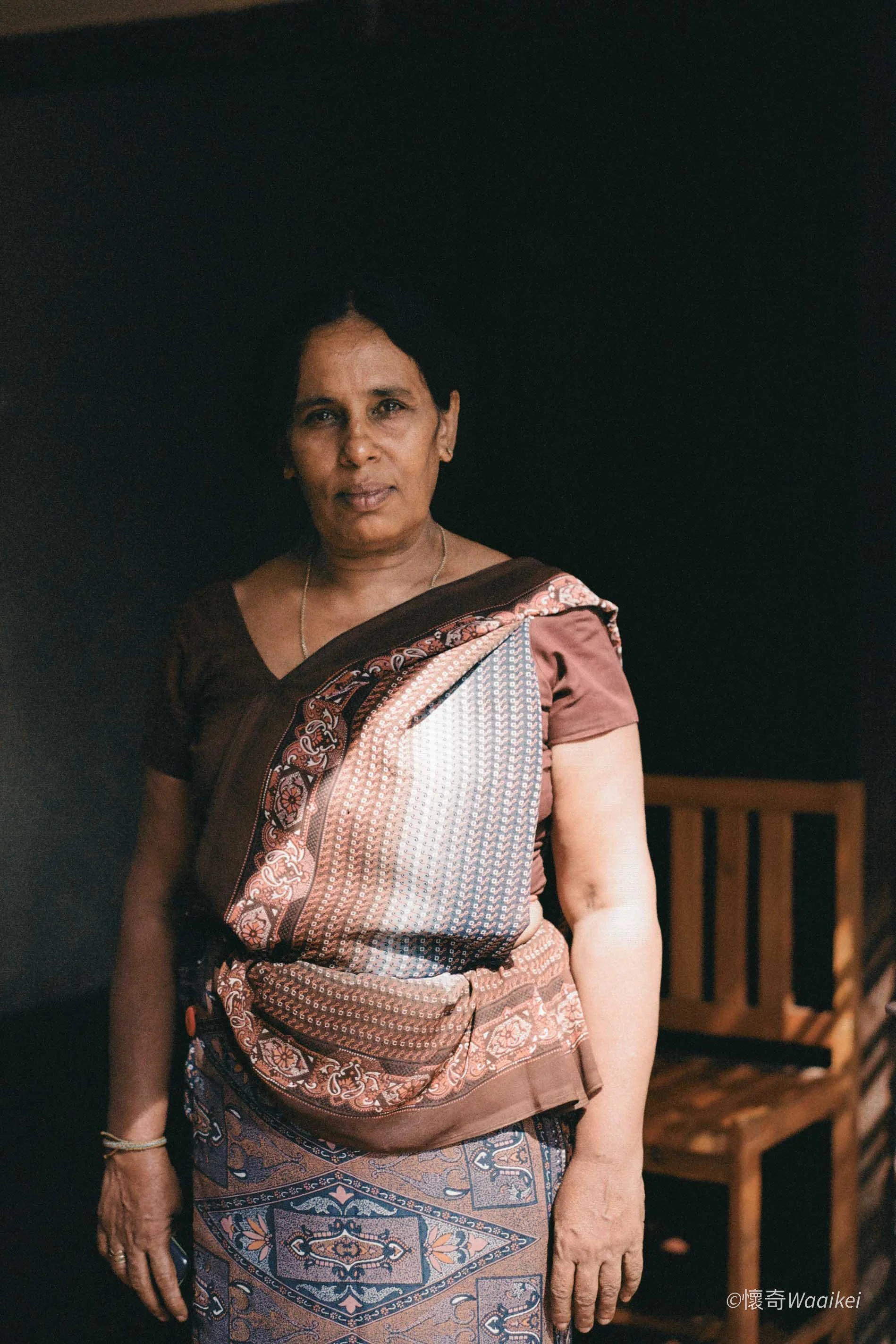 A middle-aged woman with short dark hair wearing a traditional kurta in shades of pink and maroon with intricate embroidery and patterns, standing indoors against a dark background with wooden furniture.