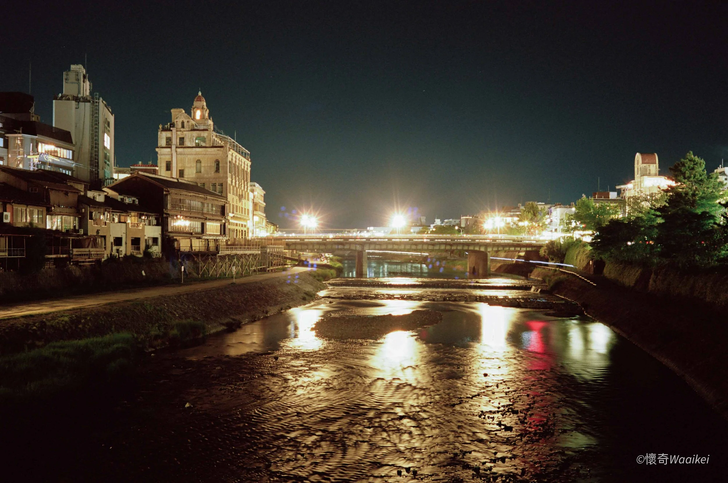 Nighttime cityscape with illuminated buildings, a bridge, and a river with reflections.