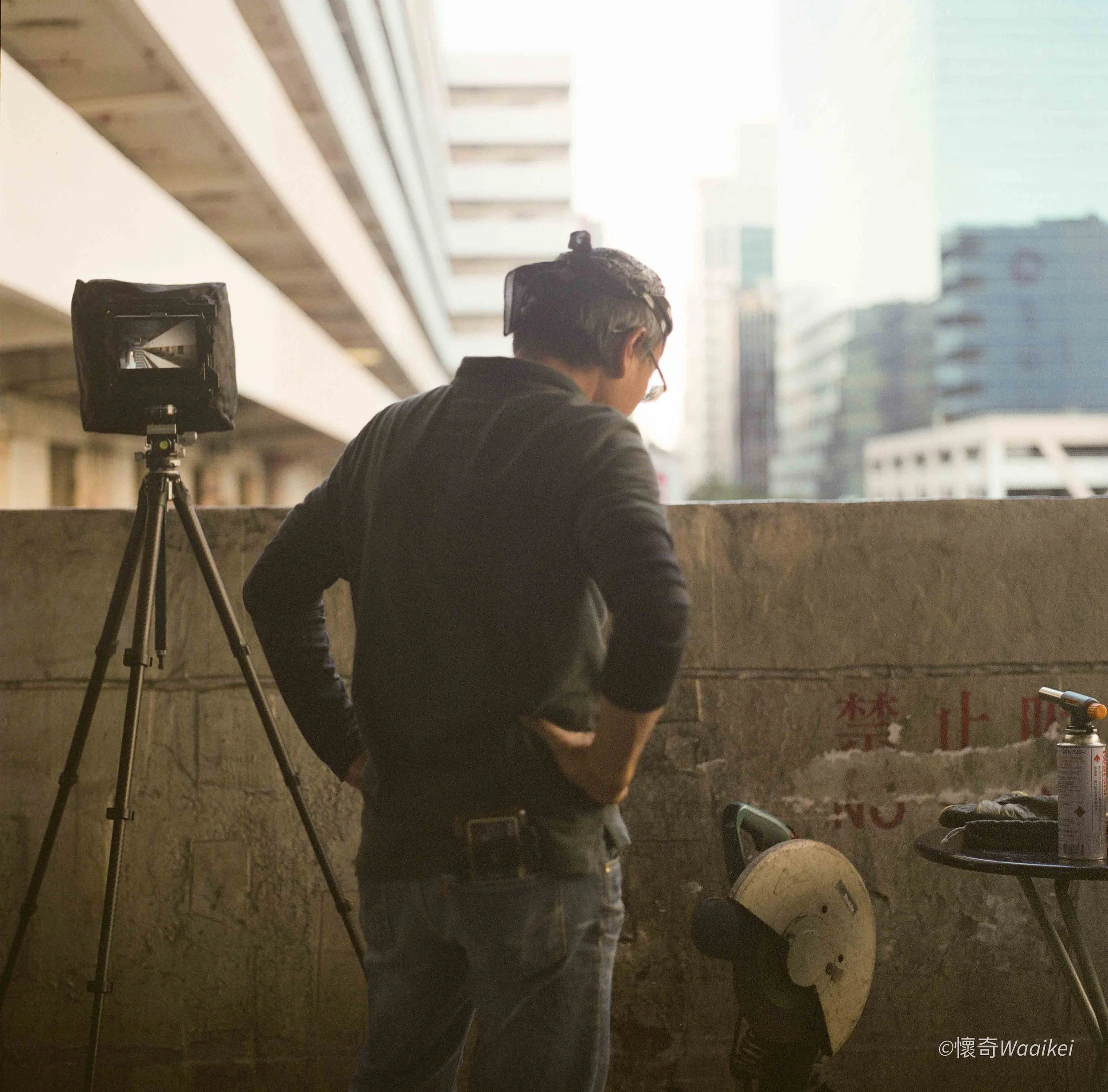 A person with gray hair and glasses, wearing a black jacket, is sitting outdoors in front of a cityscape with tall buildings. There is a camera on a tripod set up nearby, pointing towards the person. The background shows modern high-rise buildings un